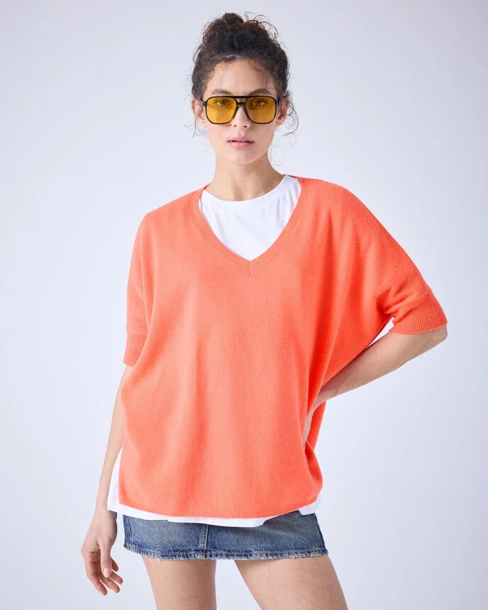 Junge Frau trägt eine weiße T-Shirt, einen roten Pullover und eine Jeansshorts, ergänzt durch Sonnenbrille mit gelbem Rahmen, stehend vor neutralem Hintergrund.