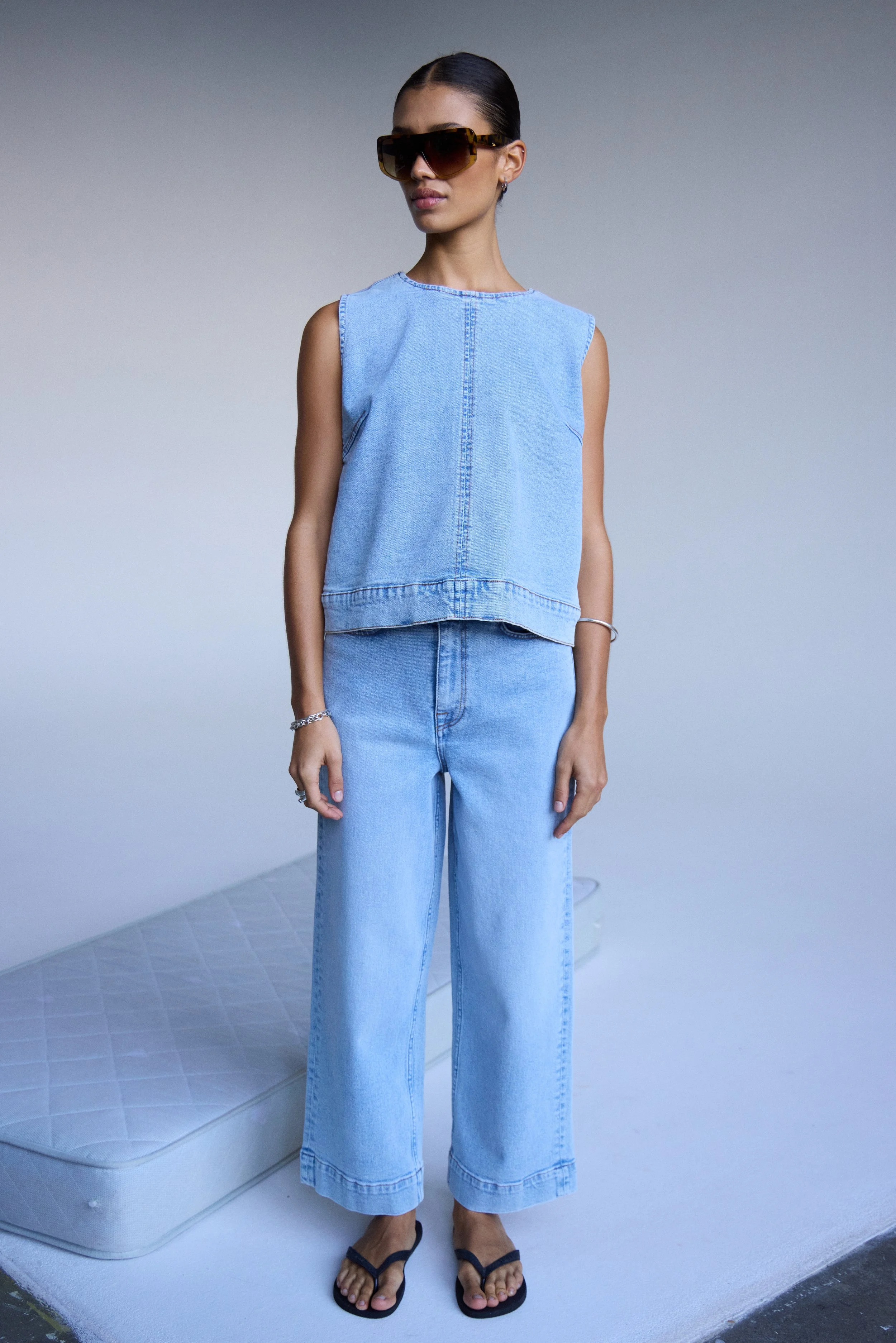 Frau trägt eine lässige Jeans- und Top-Kleidung, große Sonnenbrille, Sandalen, im minimalistisch, hellen Raum.