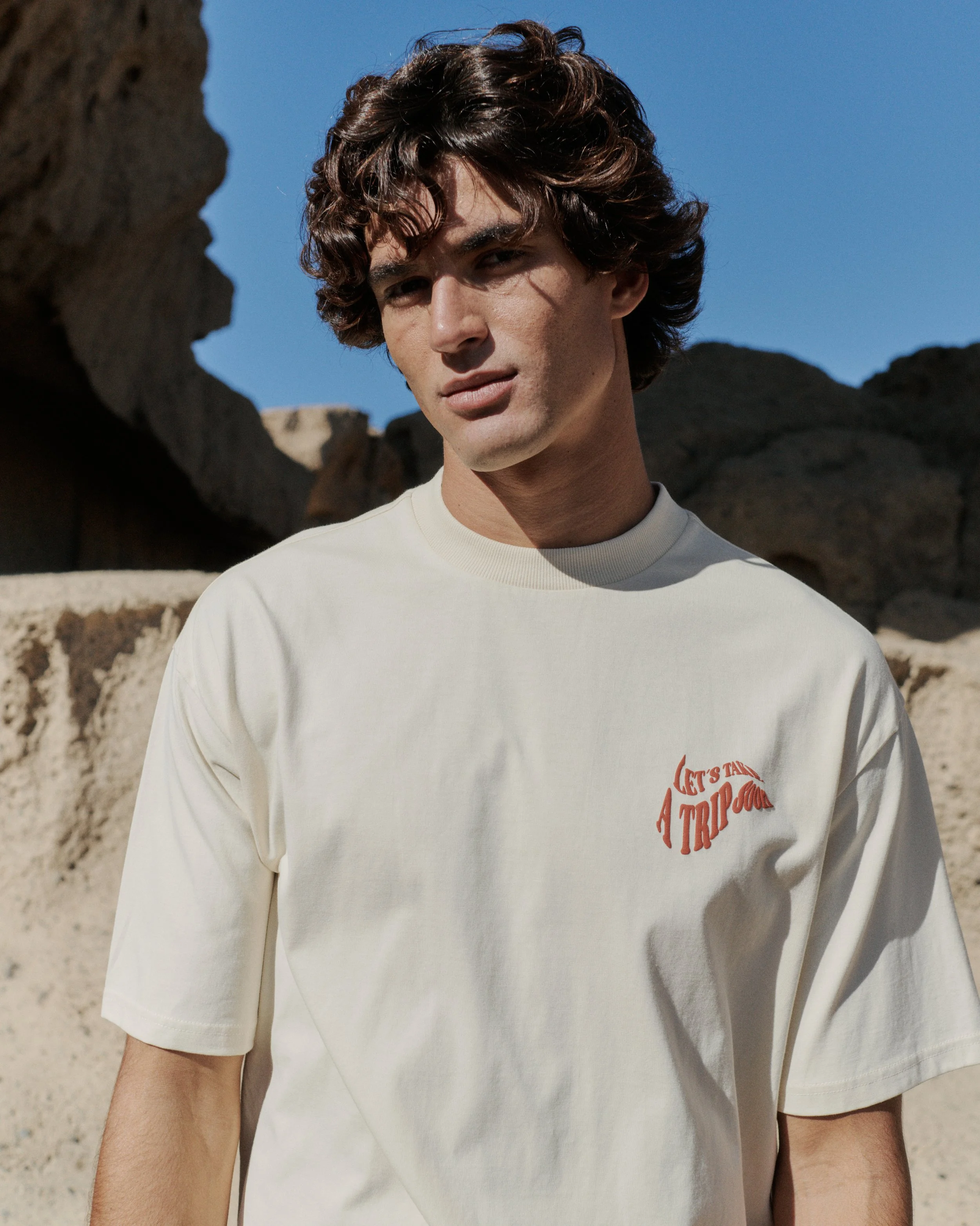 Junger Mann mit dunklen, lockigen Haaren trägt ein weißes T-Shirt mit rotem Aufdruck im Wüsten-ähnlichen Gelände bei sonnigem Himmel.