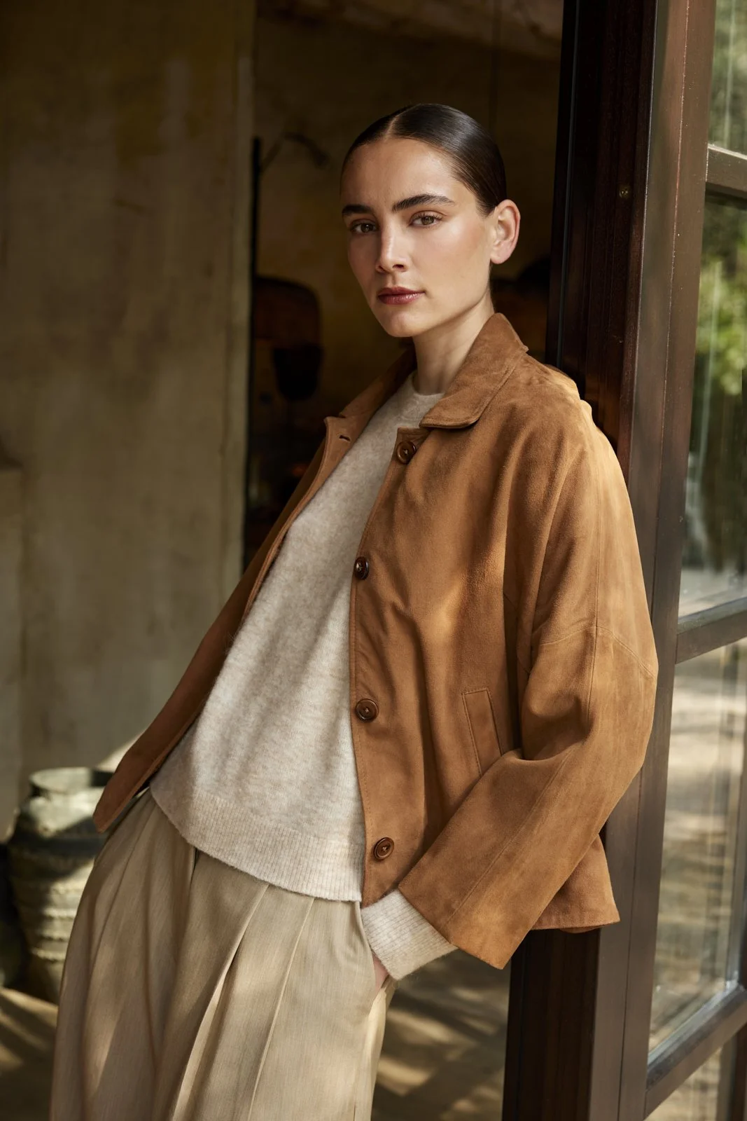 Eine junge Frau mit dunklem, glatten Haar trägt eine braune Jacke und einen hellen Pullover. Sie lehnt an einem Türrahmen neben einem Fenster.