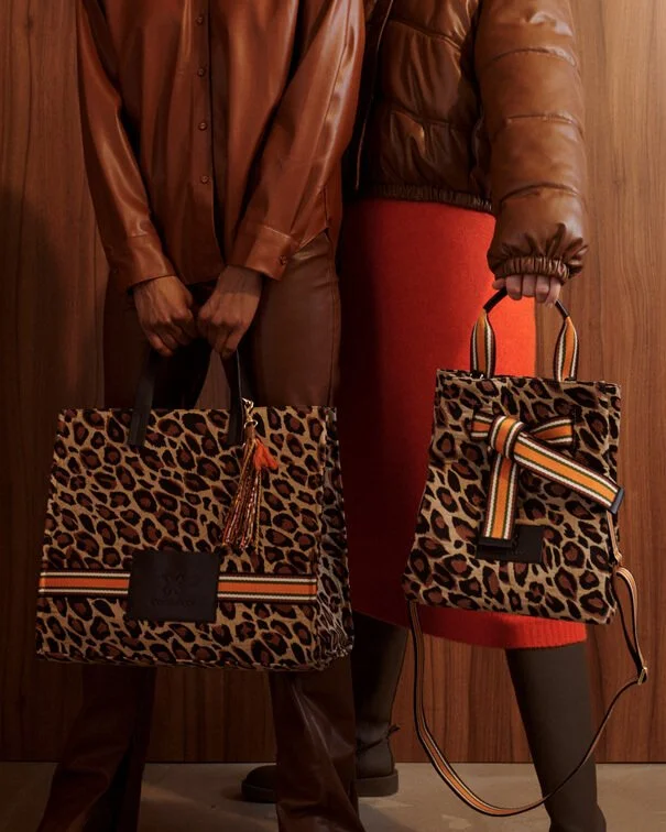 Zwei Menschen stehen nebeneinander und halten große, leopardenkopierte Taschen, die mit bunten Streifen verziert sind, vor einer Holzvertäfelung.