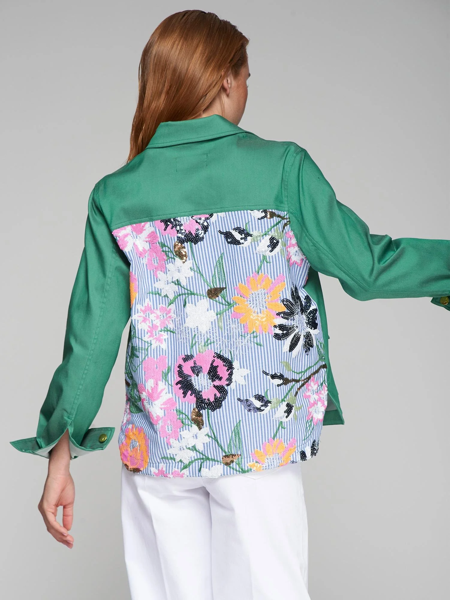 Frau trägt eine grüne Jacke mit floral besticktem Rückenteil auf einem grauen Hintergrund.