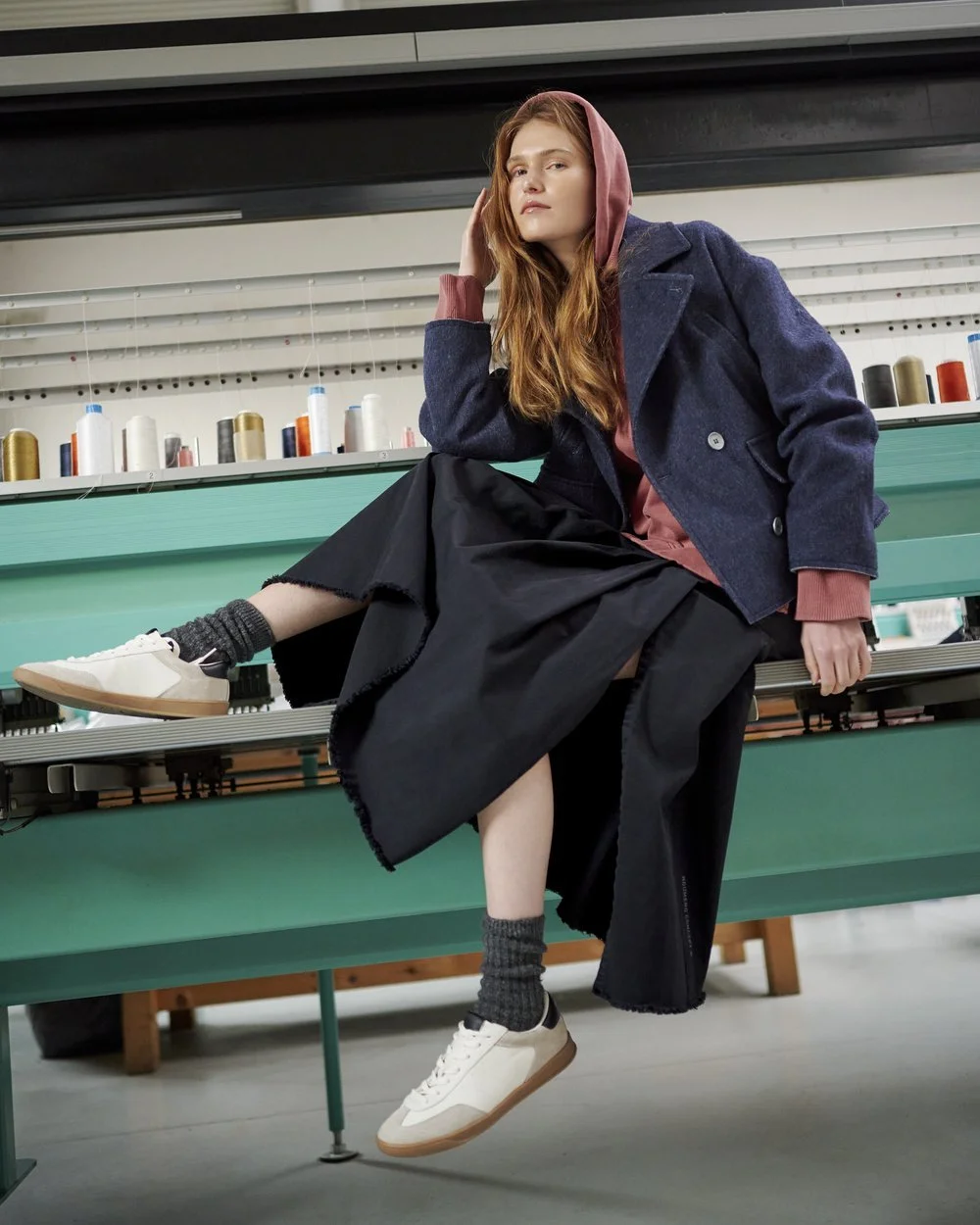 Eine junge Frau sitzt auf einer grünen Werkbank in einer Näherei mit Garnrollen im Hintergrund. Sie trägt einen rosa Hoodie, einen blauen Mantel, einen schwarzen Rock und weiße Turnschuhe mit grauen Socken. Sie hat langes, rotes Haar und schaut nach 