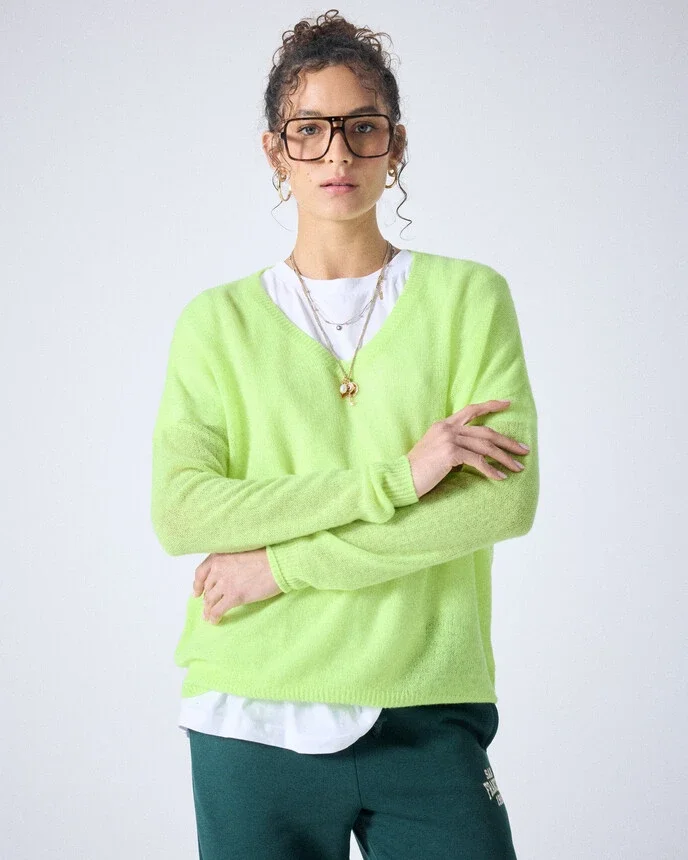 Eine junge Frau mit lockigem Haar, Brille und Ohrringen trägt eine neon grüne Strickjacke, eine weiße Bluse und dunkle Hose, steht vor hellem Hintergrund.