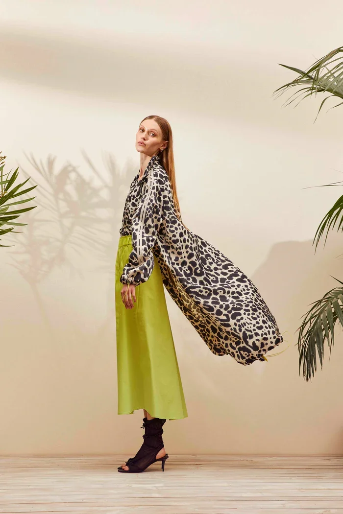 Frau in einer schwarzen, weißen und gelb gemusterten Jacke, grünen Rock, schwarzen High Heels und umhüllend eines Leopardenmusters, steht vor einer hellen Wand mit Pflanzen im Hintergrund.