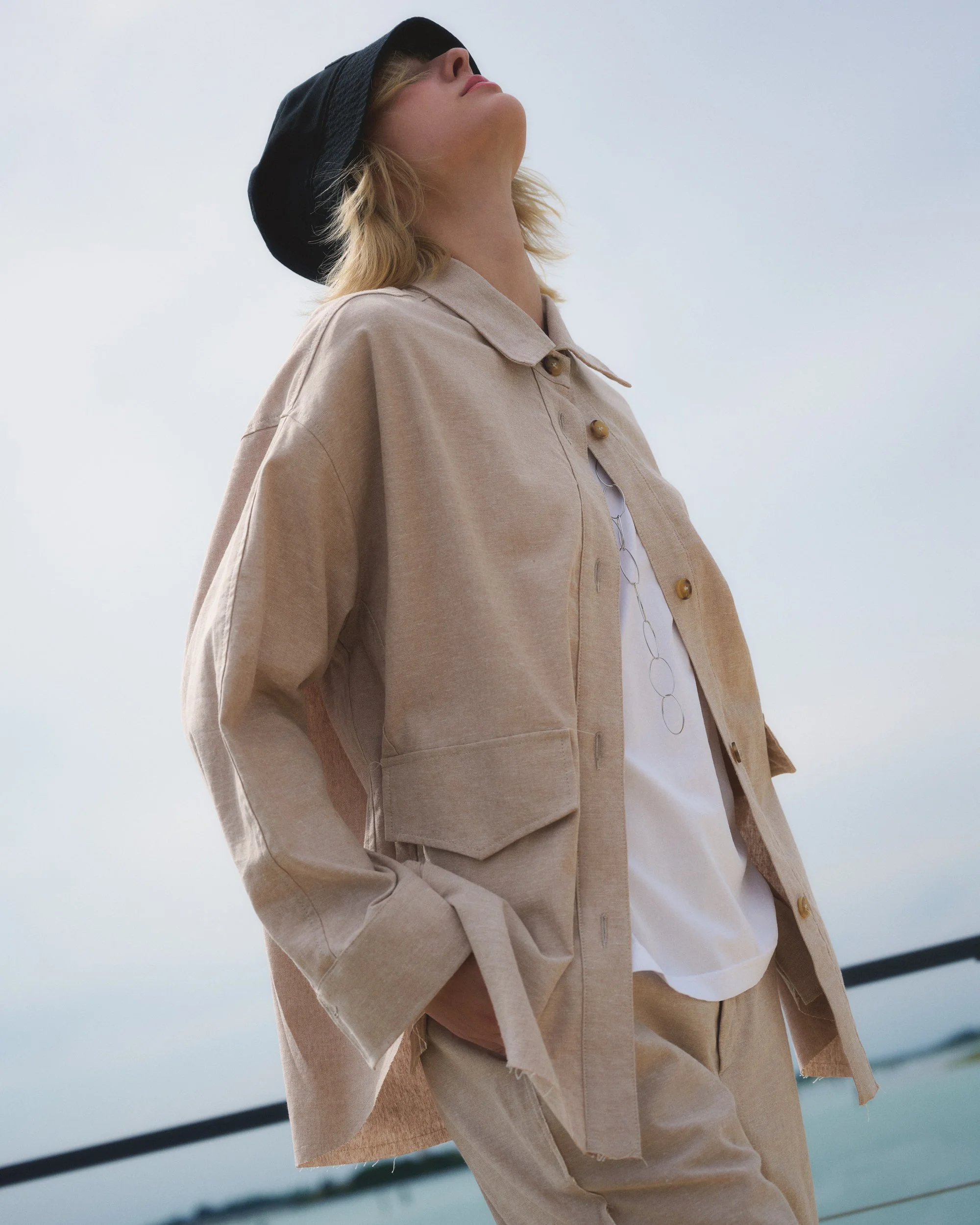 Frau mit beige Jacke, weißen T-Shirt, schwarzem Hut und Sonnenbrille blickt nach oben, vor unscharfem Hintergrund am Wasser.