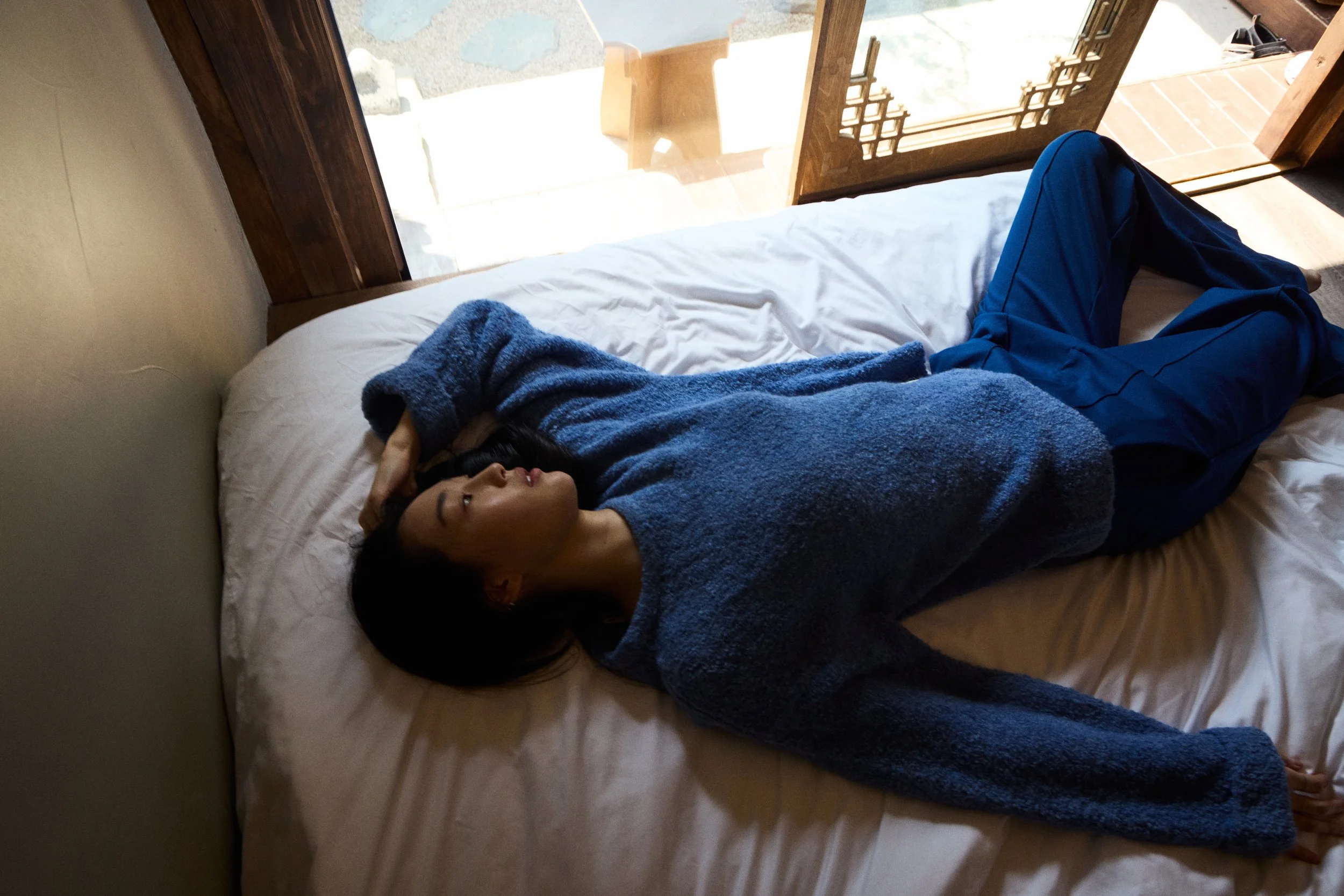 Junge Frau liegt auf einem Bett, trägt einen blauen Pullover und eine blaue Hose, mit dem Kopf zum Fenster gerichtet, in einem Raum mit Holzrahmen und Blick nach draußen.