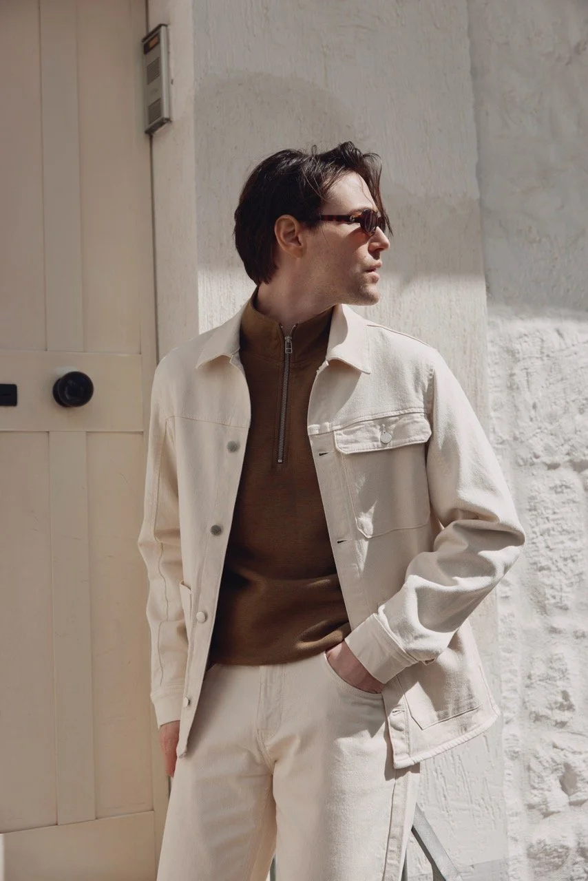 Mann trägt Sonnenbrille, beige Jacke und braunen Pullover, lehnt an einer weißen Wand im Sonnenschein.