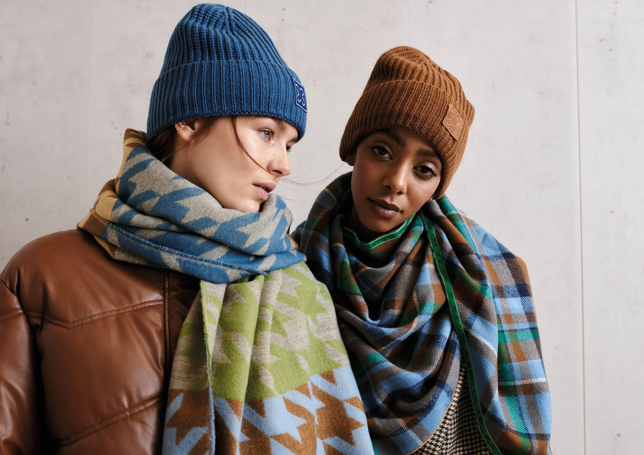 Zwei Frauen tragen Wintermützen, Schals und Jacken und stehen nebeneinander vor einer hellen Wand.