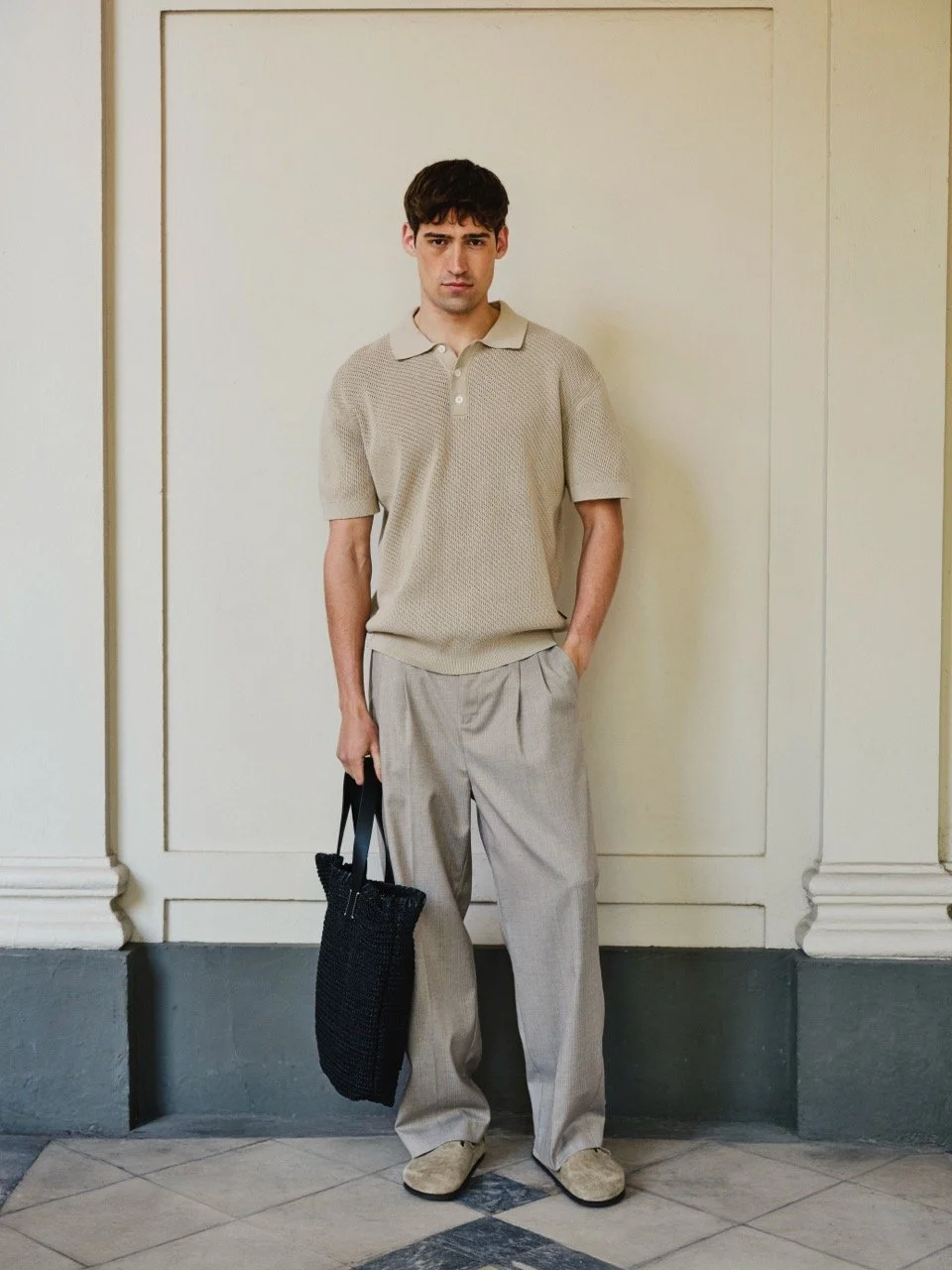 Ein Mann in beige Kleidung steht vor einer weißen Wand, hält eine schwarze Tasche, trägt beige Schuhe und schaut ernst.