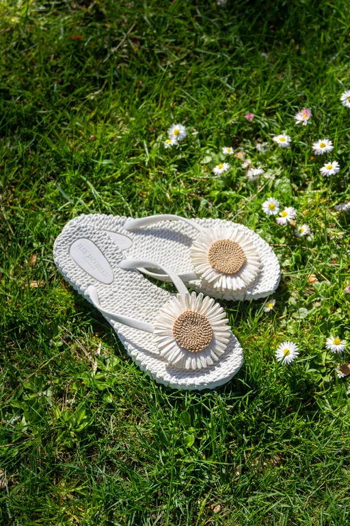 Weiße Flip-Flops mit Kamillenblumenmuster liegen auf grünem Gras mit kleinen weißen Gänseblümchen.