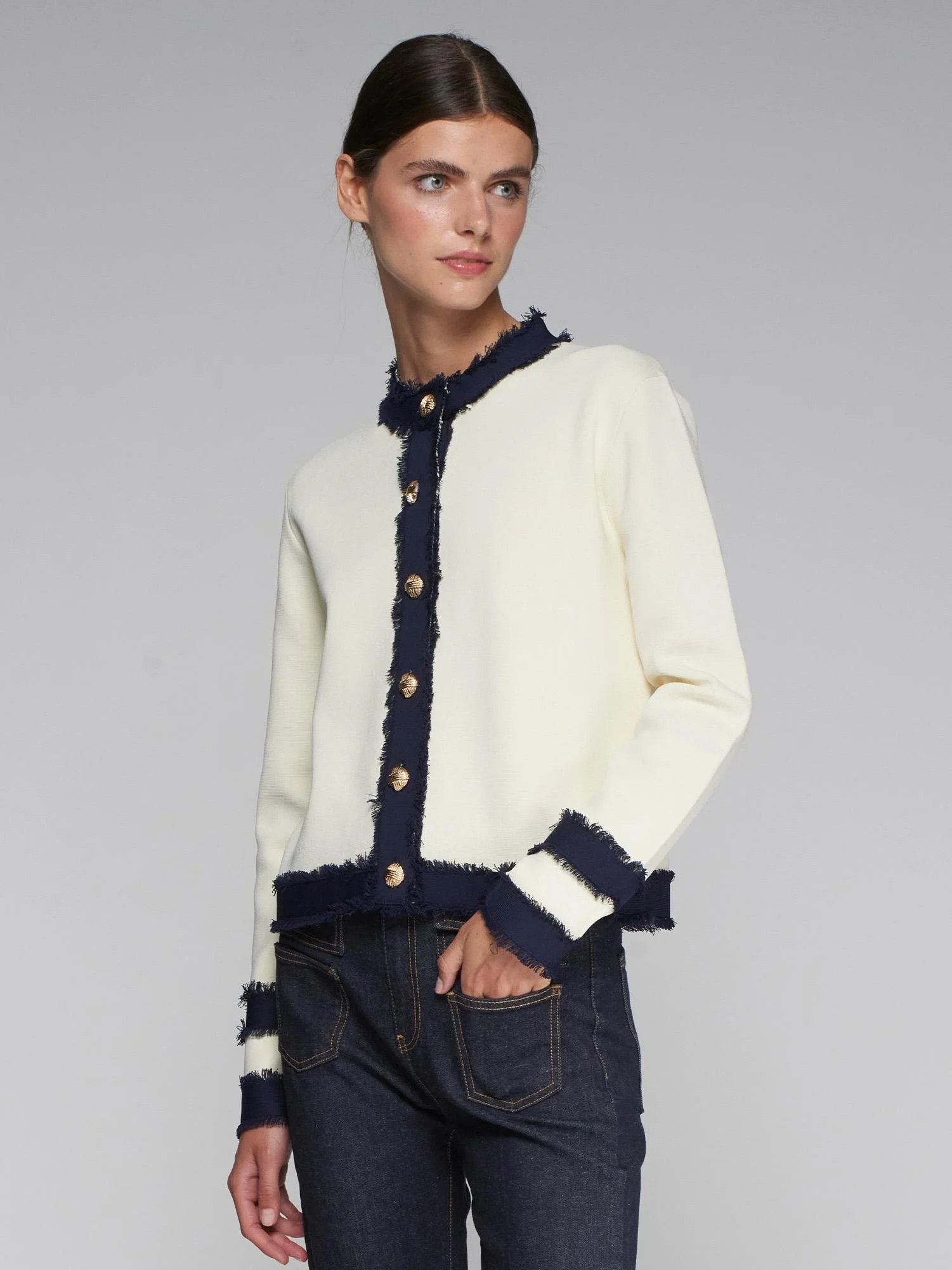 Eine junge Frau trägt eine weiße Jacke mit dunkelblauen, fransigen Akzenten und goldenen Knöpfen, kombiniert mit dunklen Jeans.