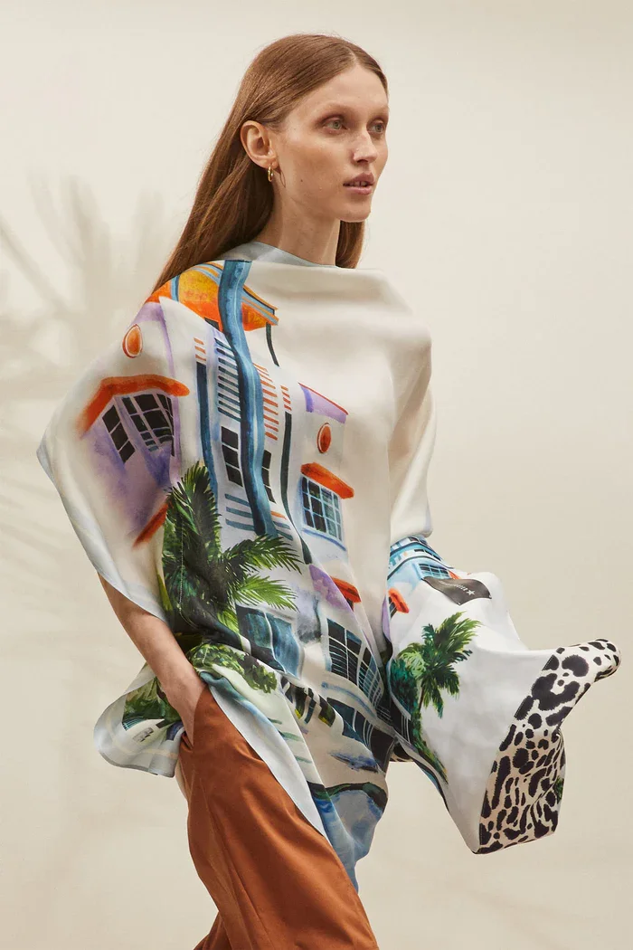 Eine Frau trägt ein kariertes Hemd, das wie ein bunter Druck eines Stadtbildes aussieht, mit Palmen und Häusern, und hat braune Hosen an.