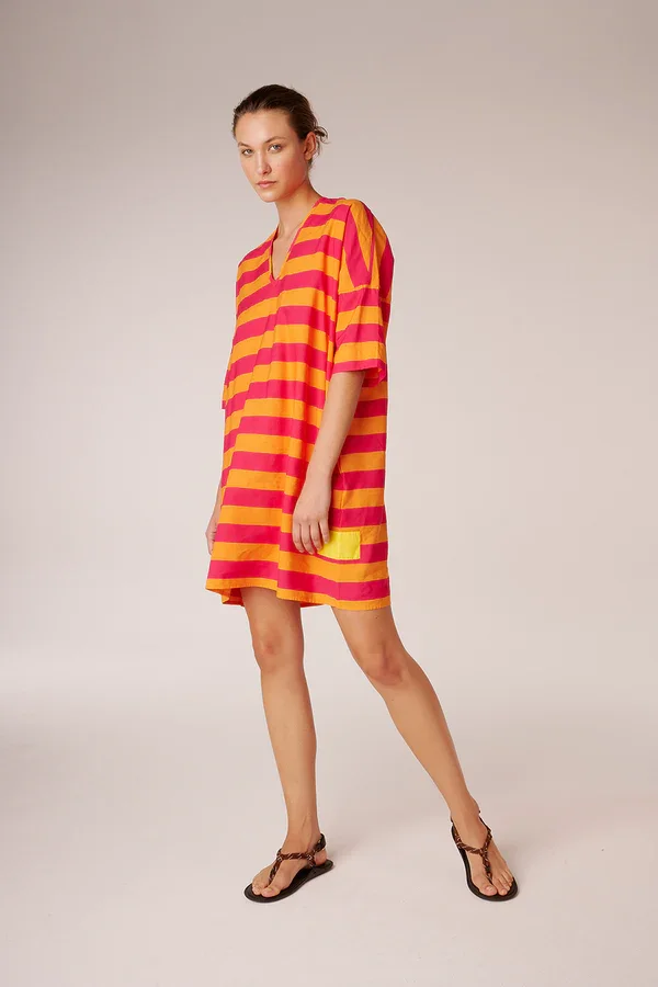 Frau trägt ein rotes und gelbes gestreiftes Strandkleid, steht aufrecht vor neutralem Hintergrund, trägt braune Sandalen.