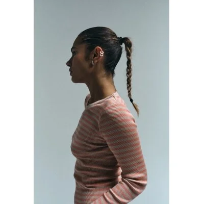 Frau mit gestreiftem pinken Pullover und geflochtenem Zopf, seitliche Ansicht, dunkle Haare, neutraler Hintergrund.