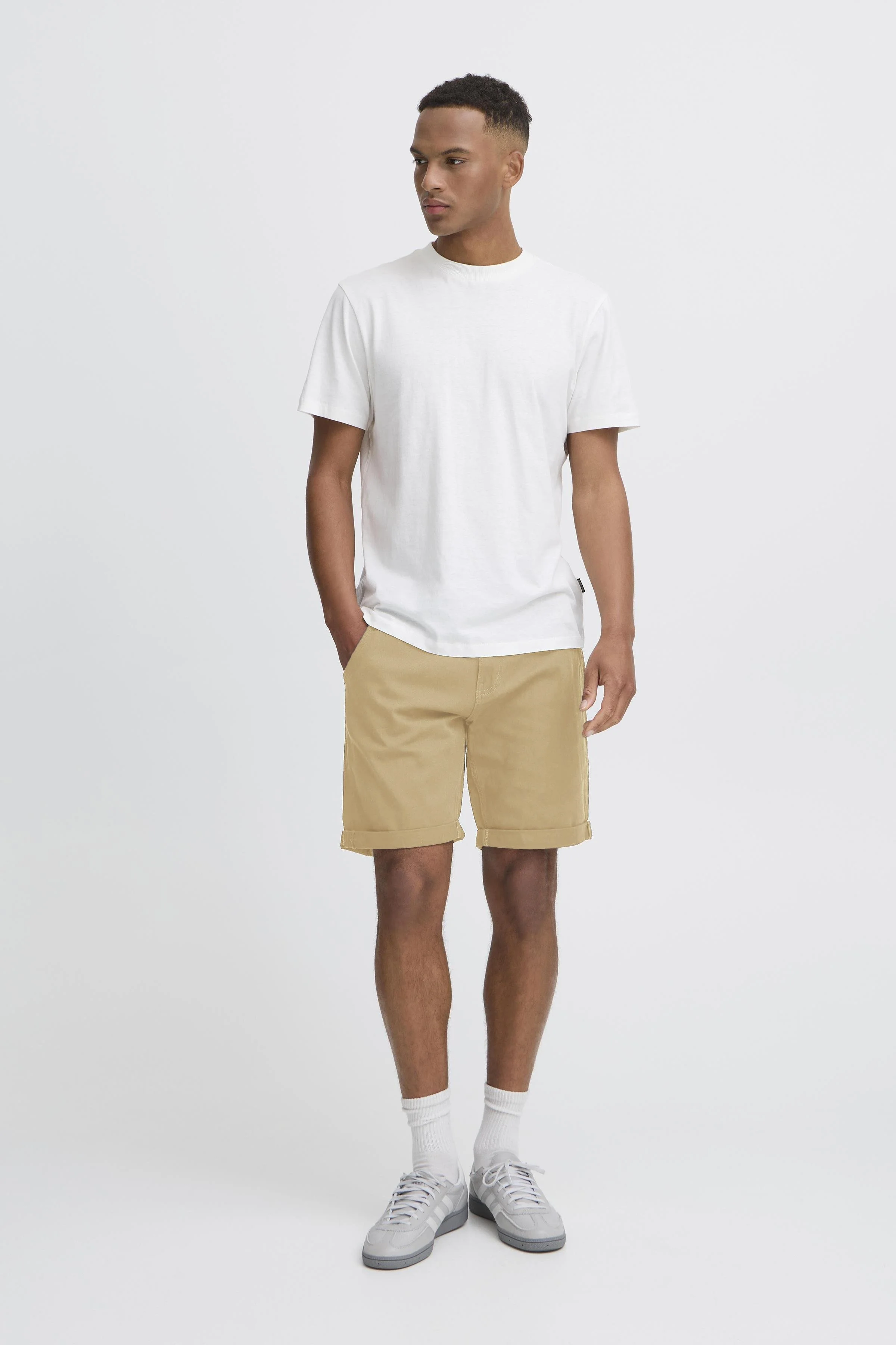 Mann trägt weißen T-Shirt, beige Shorts, weiße Socken und Sneakers, steht vor weißem Hintergrund.