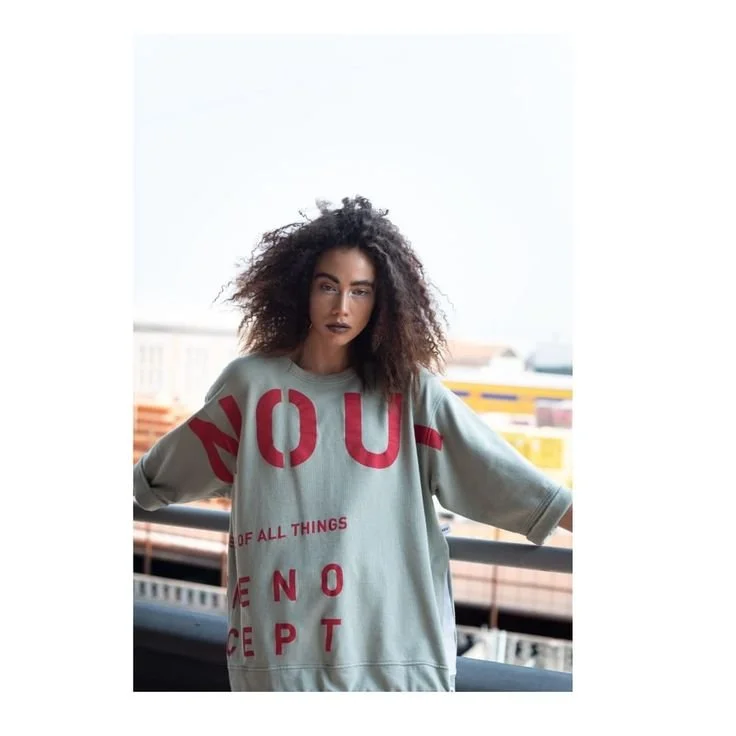 Eine Frau mit lockigem Haar posiert auf einer Stadtbalkon durch eine Kleidung mit großen roten Buchstaben, die eine Botschaft vermittelt.