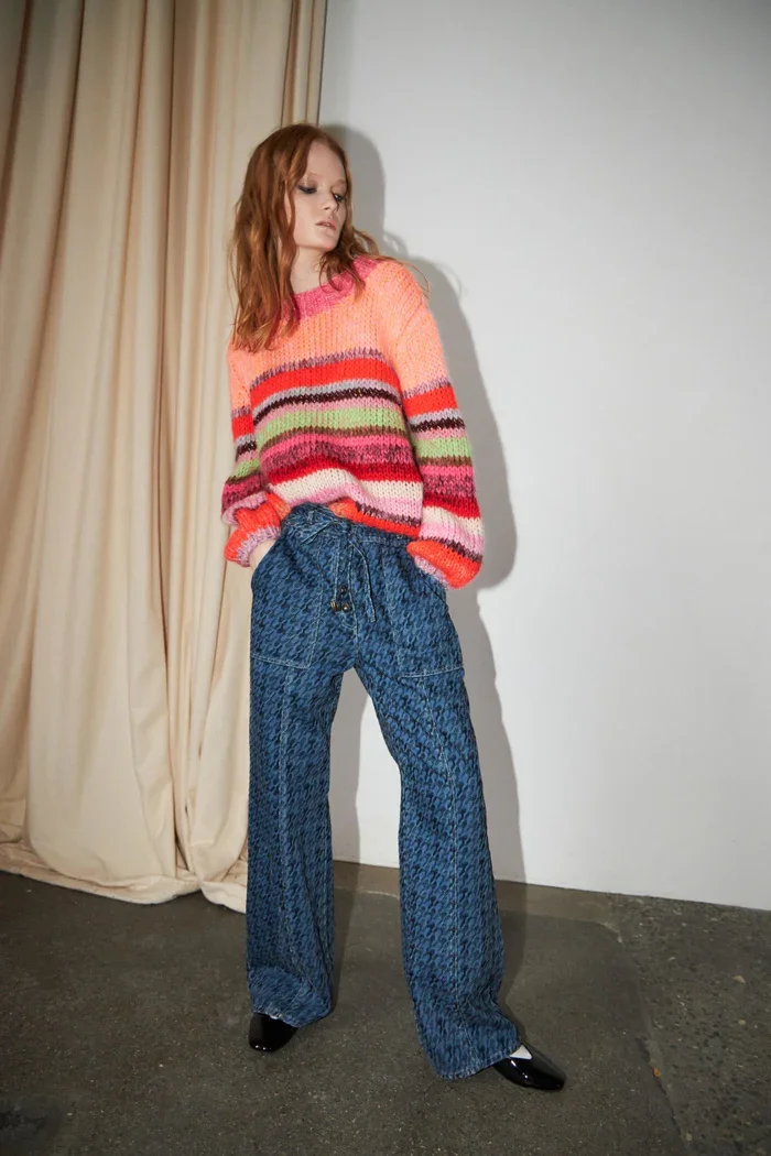 Frau steht vor einer weißen Wand, trägt eine bunte gestreifte Strickjacke, weite blaue Hosen und schwarze Schuhe.