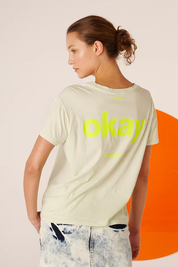 Junge Frau trägt ein cremefarbenes T-Shirt mit dem Wort 'okay' in leuchtend gelber Schrift, steht vor einem weißen Hintergrund mit orangefarbenem Kreis im Hintergrund.