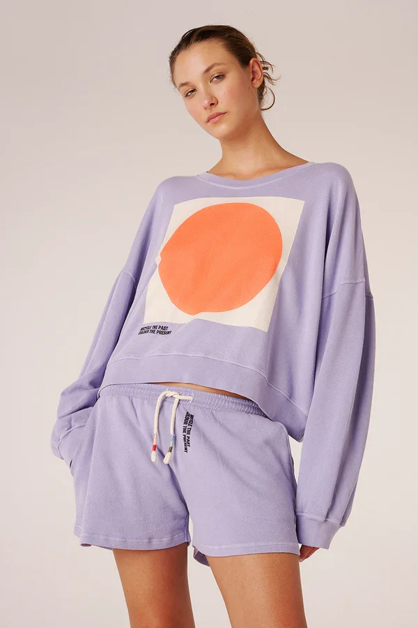 Frau trägt lilafarbenes Sweatshirt mit orangefarbenem Kreis und Shorts in derselben Farbe.