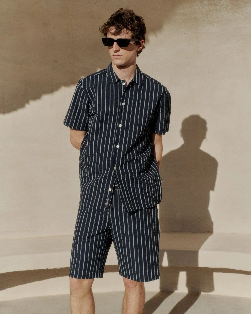 Junge Mann trägt dunkelblau-weiß gestreiftes Hemd und Shorts, trägt schwarze Sonnenbrille, steht vor heller Wand, Schatten im Hintergrund.