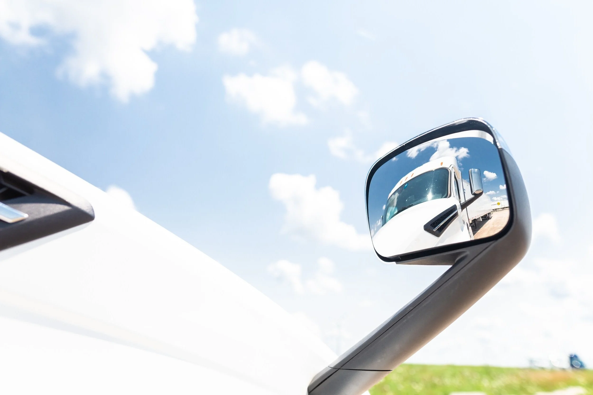 Side mirror of a white vehicle reflecting a blue sky with white clouds and a distant truck on the road.