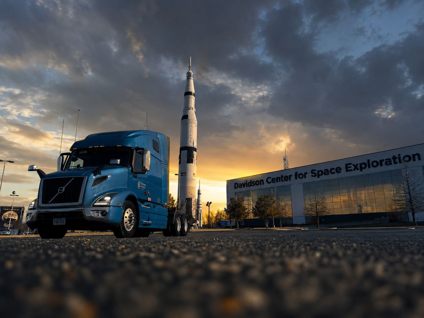 A blue MCK Trucking truck parked in front of a rocket on the launch pad at the Davidson Center for Space Exploration during sunset.