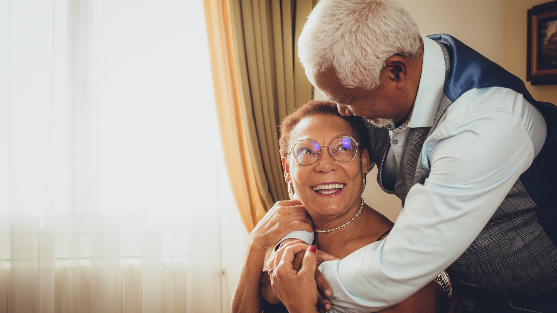 An elderly woman with glasses and a pearl necklace smiling happily as a man embraces her warmly, in a room with sunlight coming through the window.