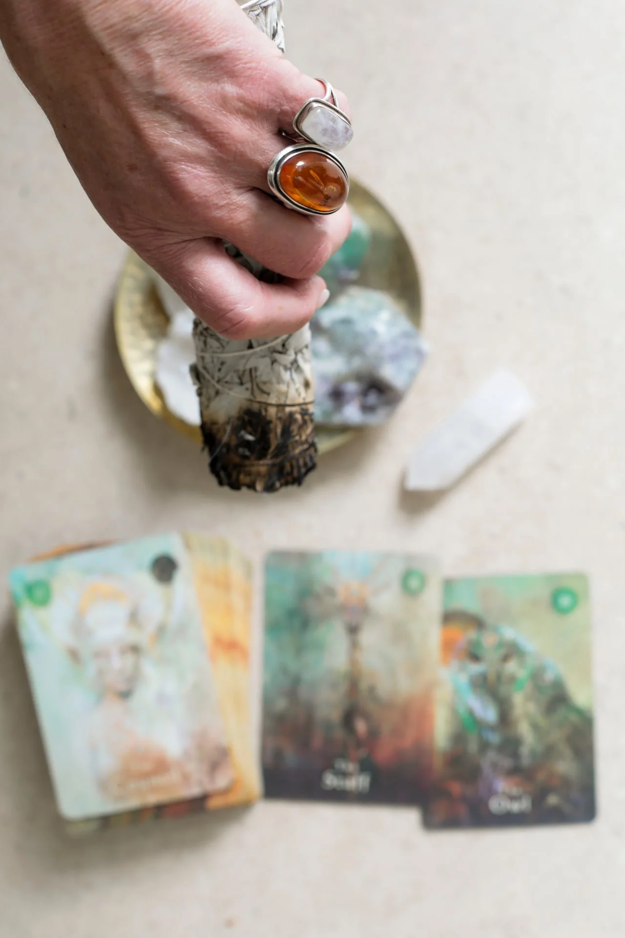 A person's hand with rings holding a bundle of sage, surrounded by crystal and colorful tarot cards on a beige surface.