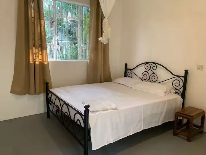 A bedroom with a metal bed frame, white bedding, a small wooden side table, a window with bars, and beige curtains.