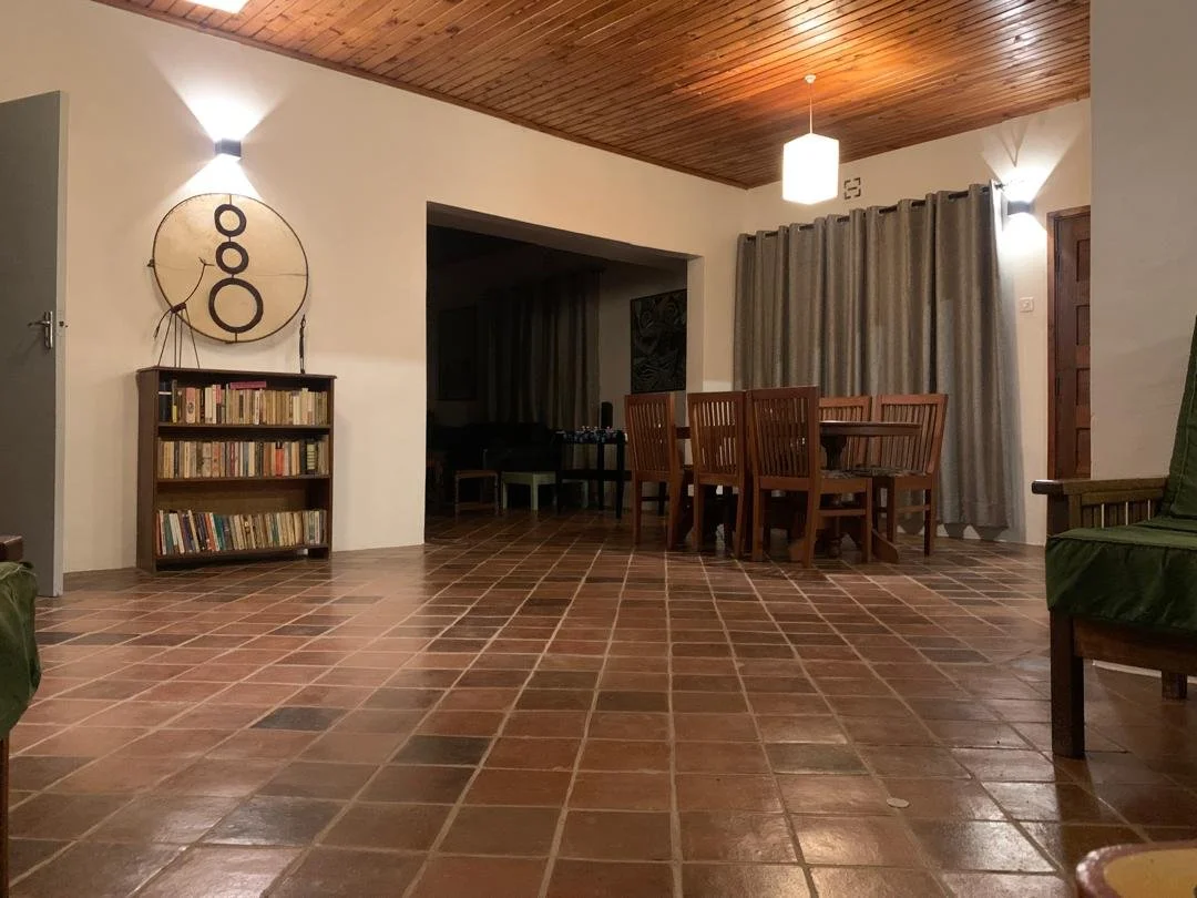 Our living room with terracotta tile floor, wooden ceiling, dining table with six chairs, bookshelf with books, and curtains covering windows.