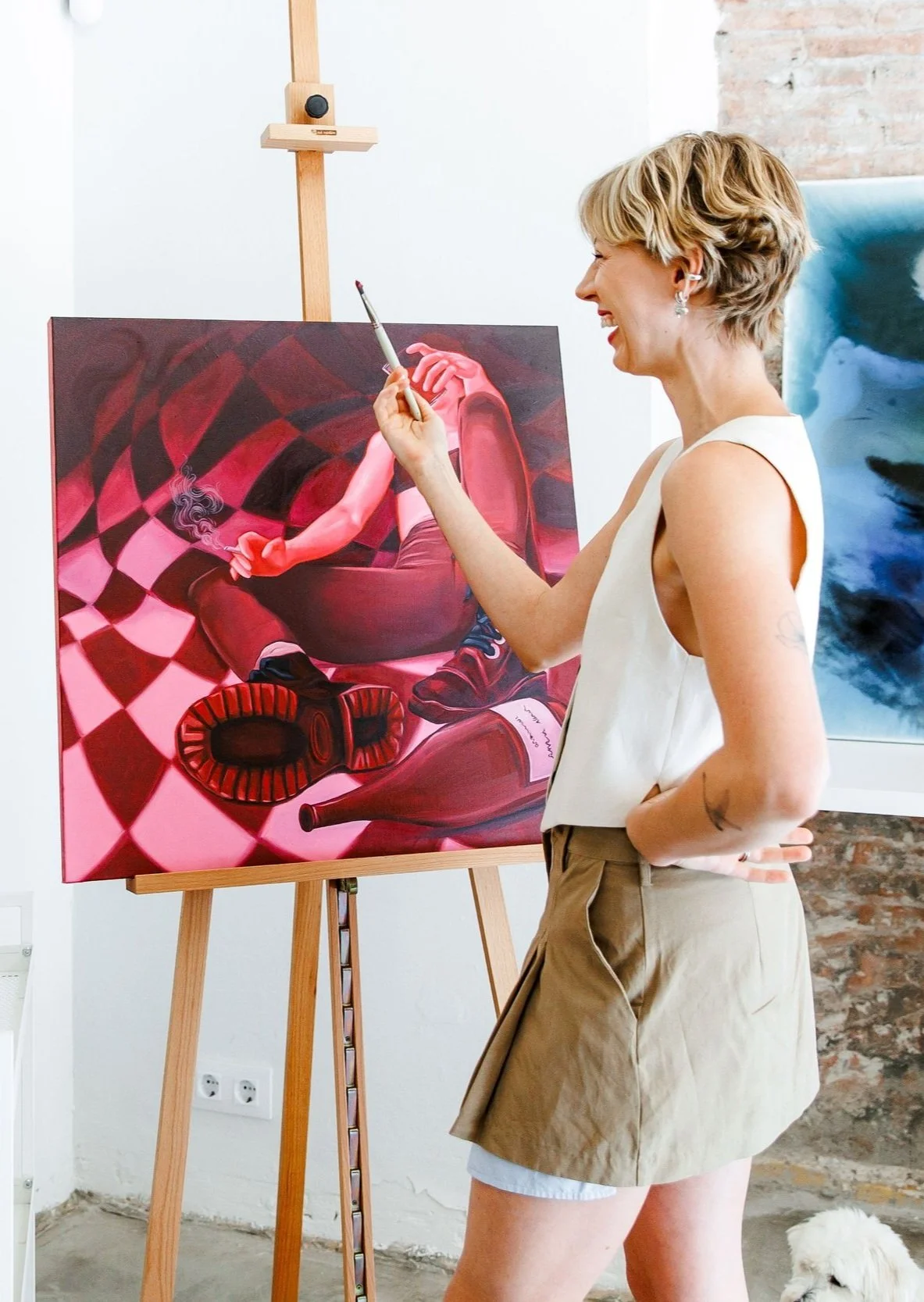 A woman with short blond hair, wearing a white sleeveless top and beige shorts, smiling as she paints a surreal artwork of a person smoking, with a tilted bottle and a checkered floor background, on a canvas in an art studio with brick and white walls.