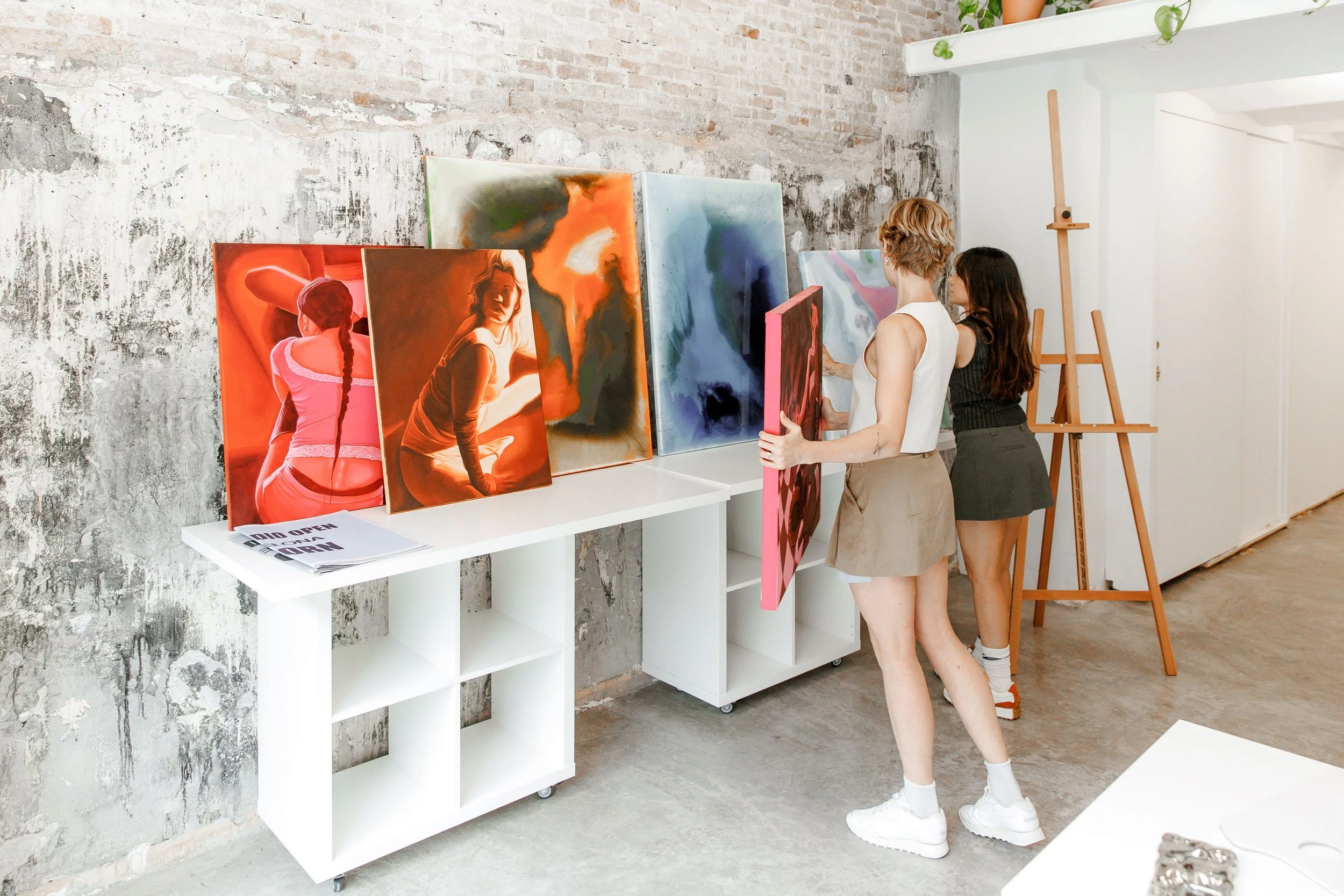 Two women viewing colorful abstract paintings displayed on white shelves in an art gallery or studio with an exposed brick wall and wooden easels in the background.