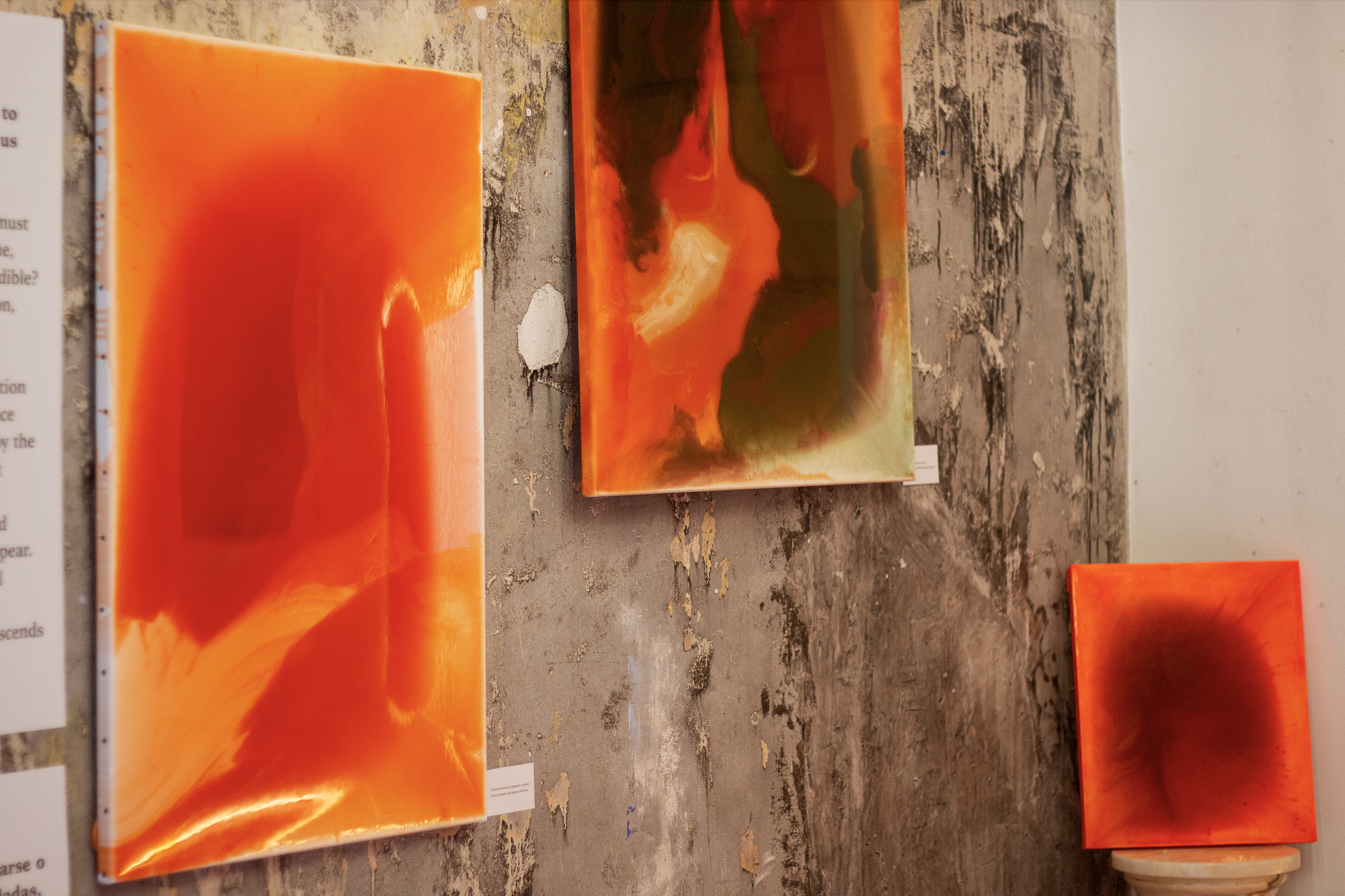 Three colorful abstract paintings with fiery orange, red, yellow, and green hues hanging on a textured wall in an art gallery.