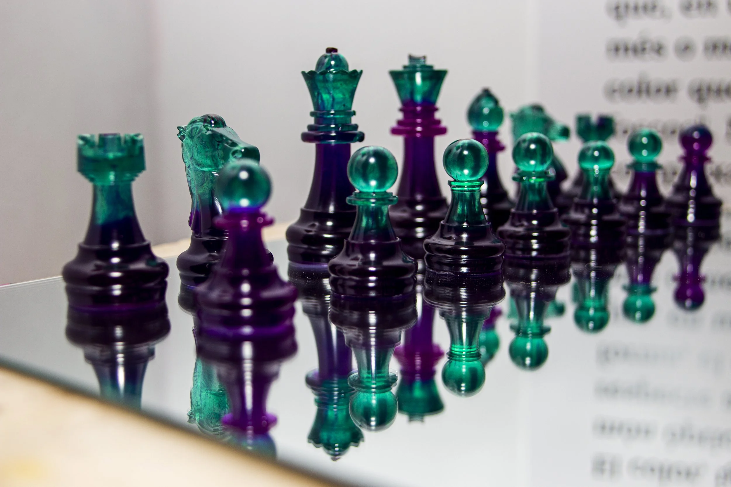 Glass chess pieces in shades of teal and purple, reflected on a glass surface, with blurred text in the background.