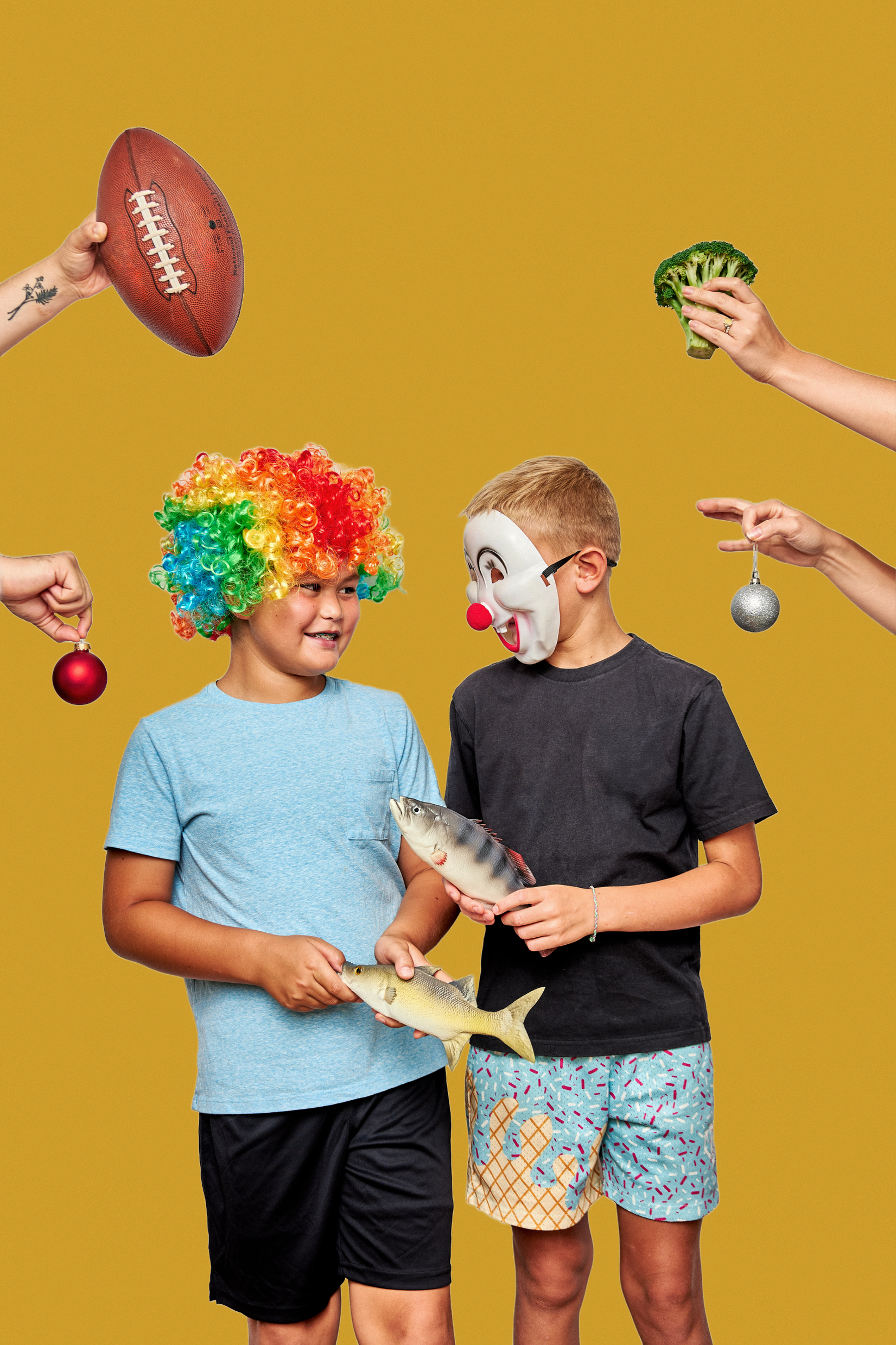 Two children standing against a yellow background, with various objects held around them. One child is wearing a rainbow-colored clown wig, and the other is wearing a clown mask. The children are holding fish, and hands around them are holding toys and objects such as a football, broccoli, Christmas ornaments, and a silver ball, creating a playful and whimsical scene.