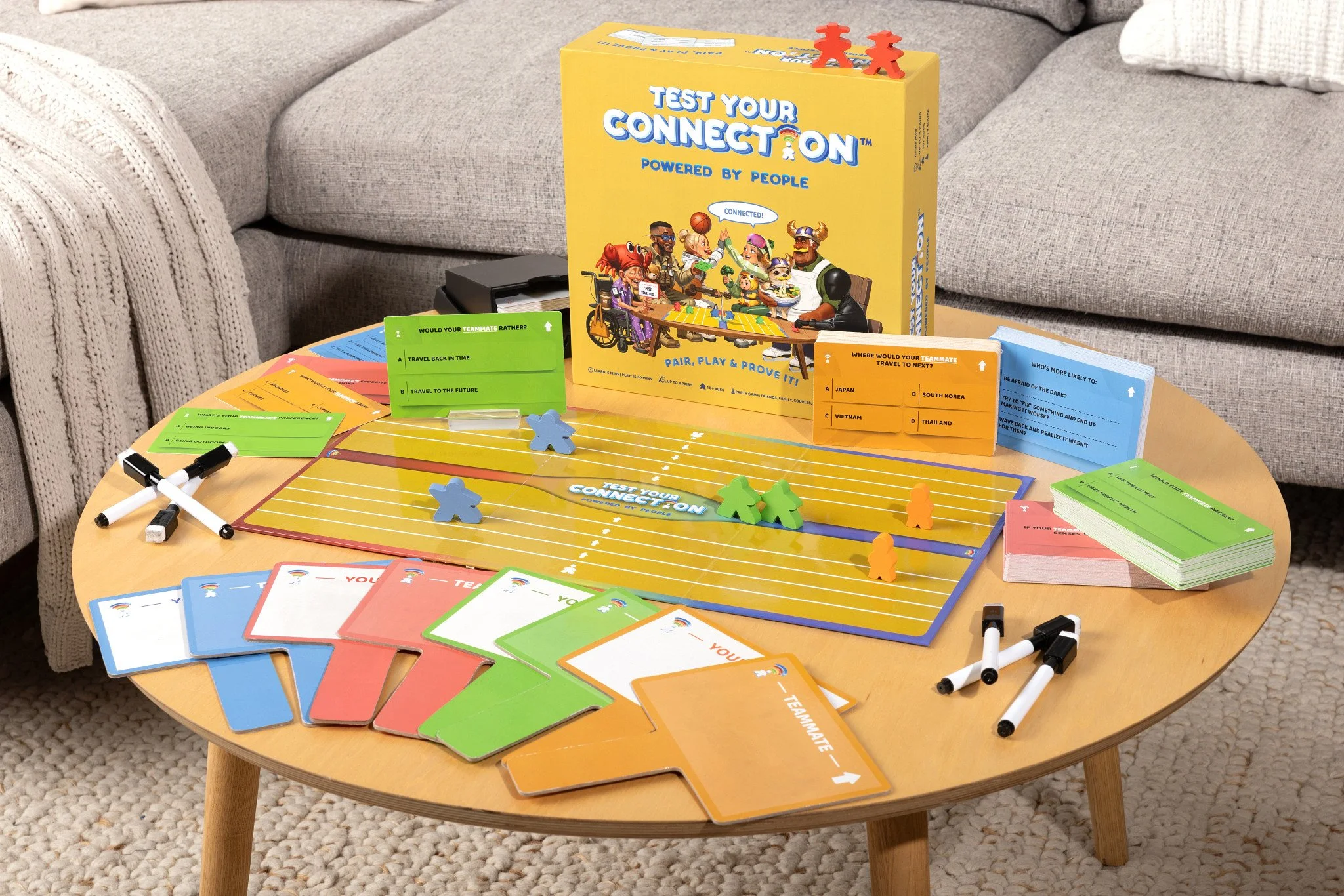 A tabletop game called 'Test Your Connection' with colorful cards, game pieces, and a yellow game box on a wooden table in a living room. The game involves matching pairs and proving connections among players.