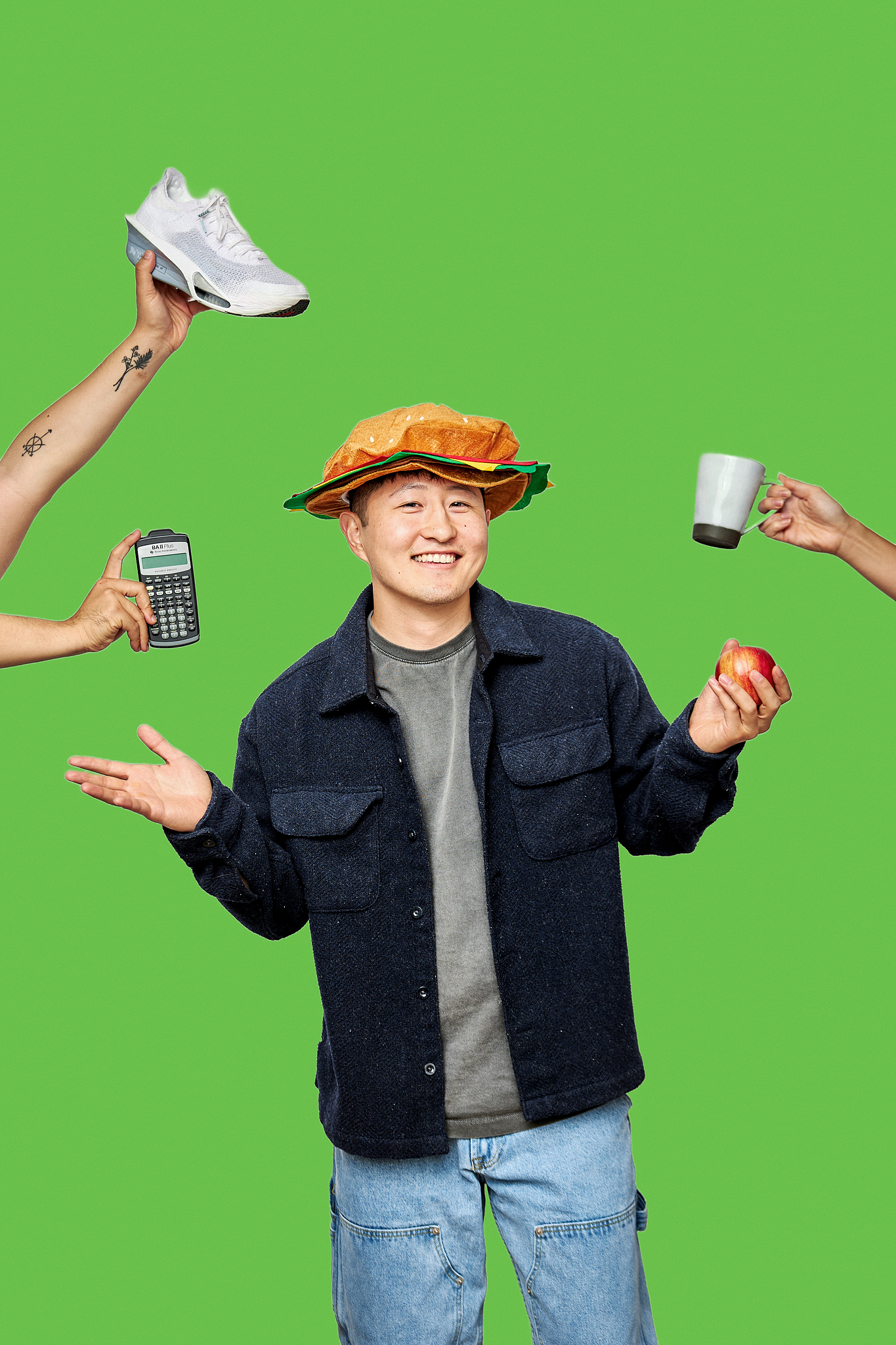 Young man smiling, wearing a hamburger hat, holding an apple, with a white sneaker, calculator, coffee mug, and tattoos on his arms against a green background.