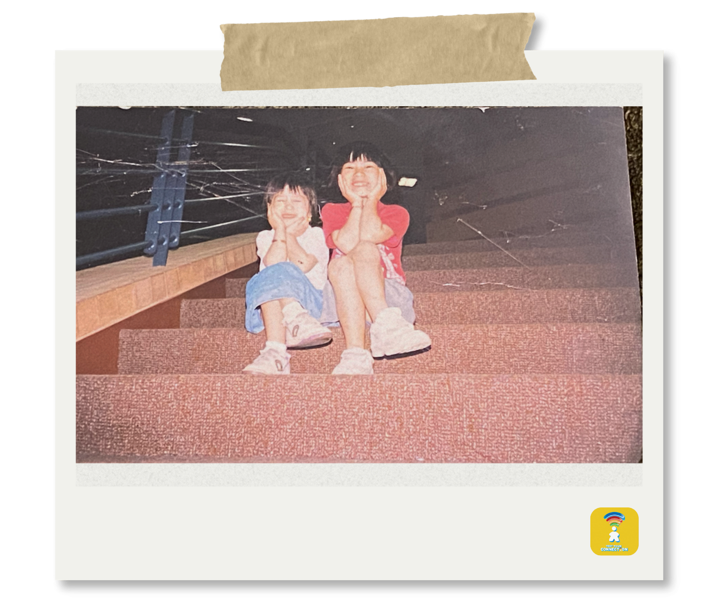 Two young girls sitting on carpeted stairs, smiling and resting their chins on their hands, with a black railing and some wiring visible in the background.