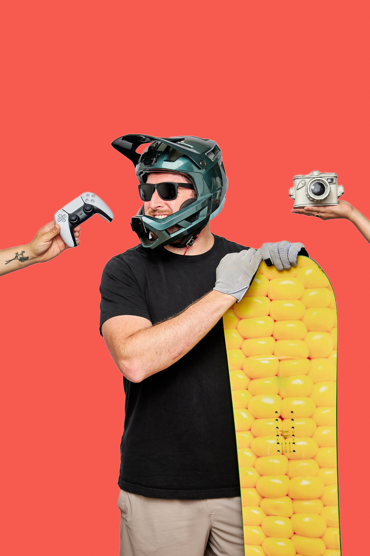 A man wearing a motocross helmet and sunglasses holding a skateboard with a yellow pattern, standing against a red background, with a hand holding a vintage camera and another holding a game controller in the frame.