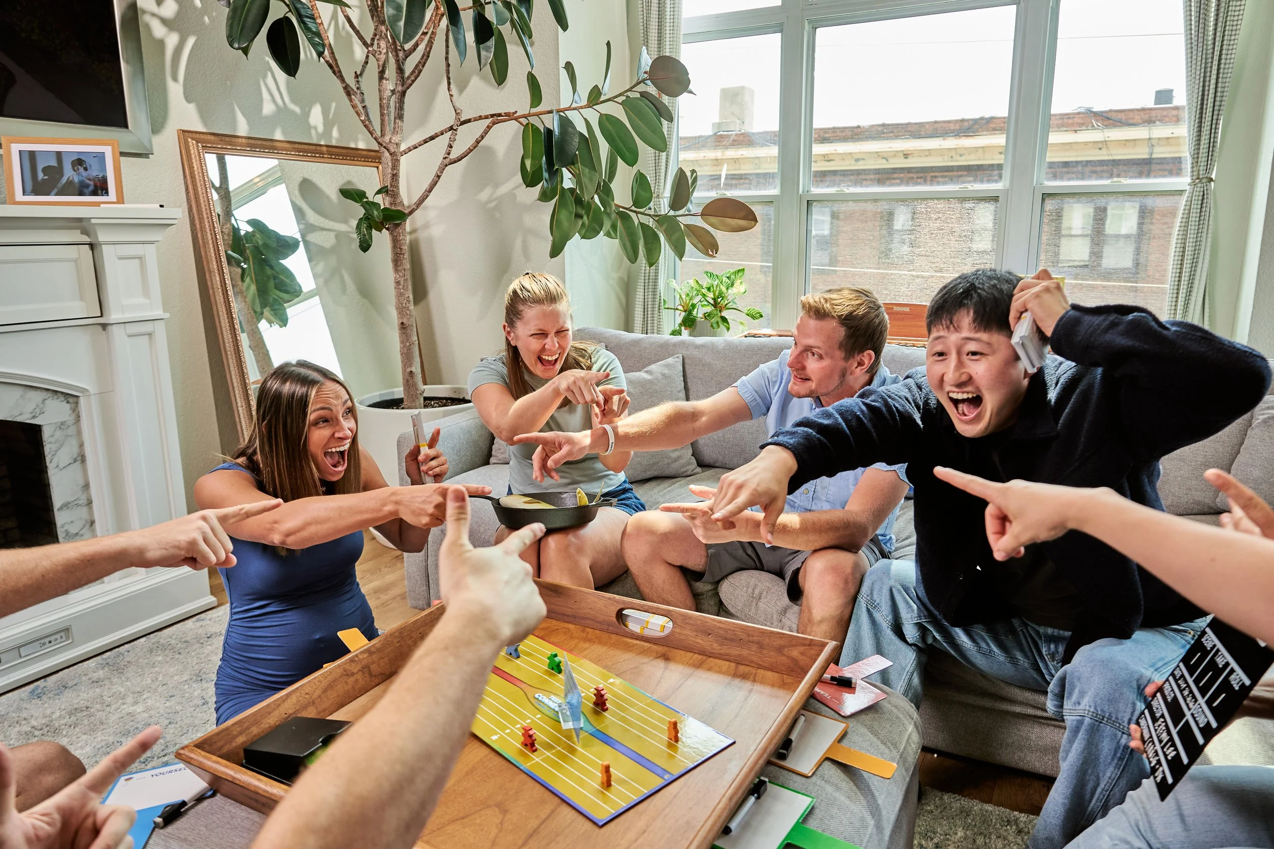 A group of people playing a tabletop hockey game, laughing and pointing at the game. They are sitting in a living room with large windows, a potted plant, and a mirror on the wall.