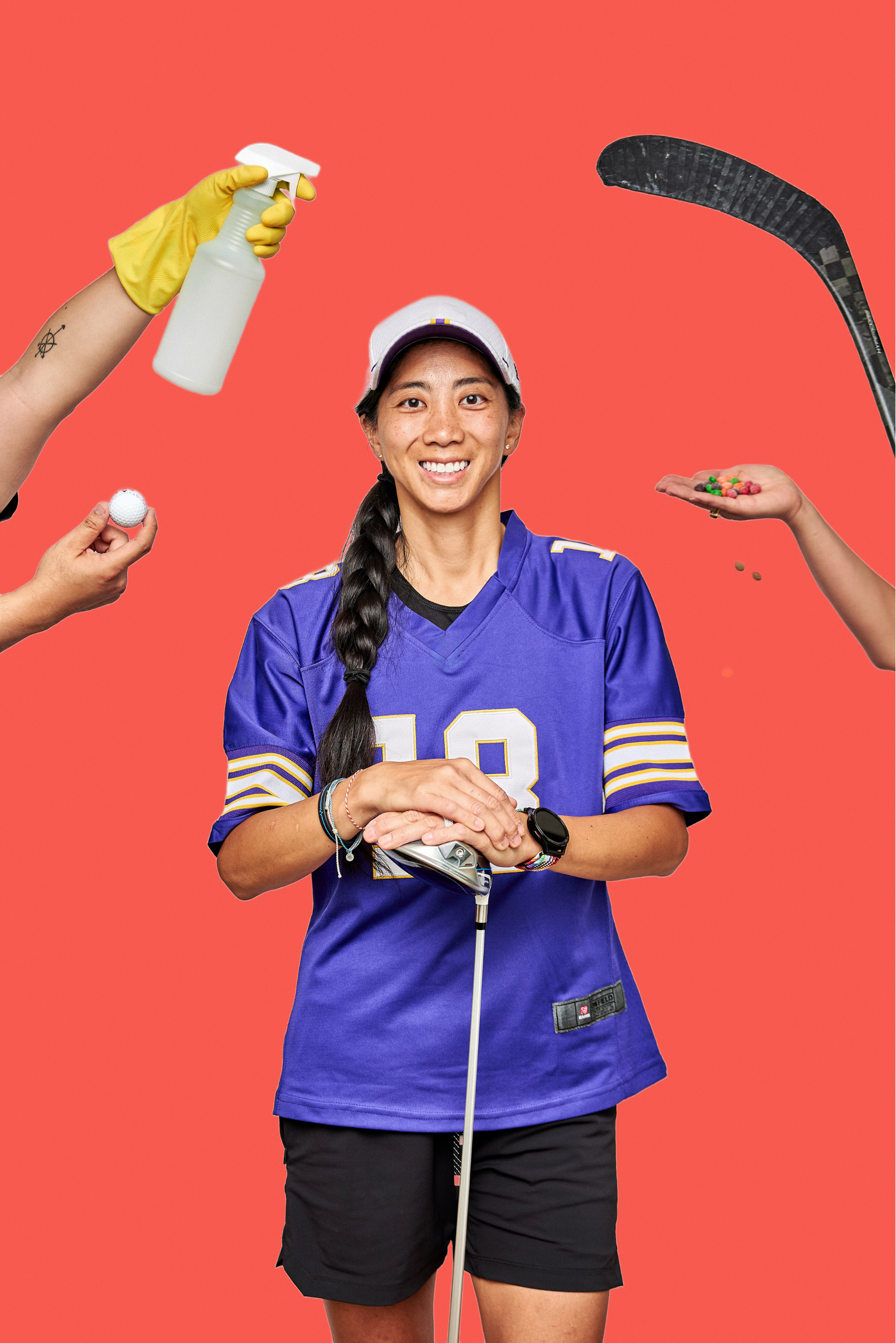 A woman wearing a purple sports jersey and black shorts is smiling and holding a golf club. She has a long braid and wears a white cap. Several hands around her are presenting various objects including a golf ball, a spray bottle, colorful candies, and a hockey stick, against a coral pink background.