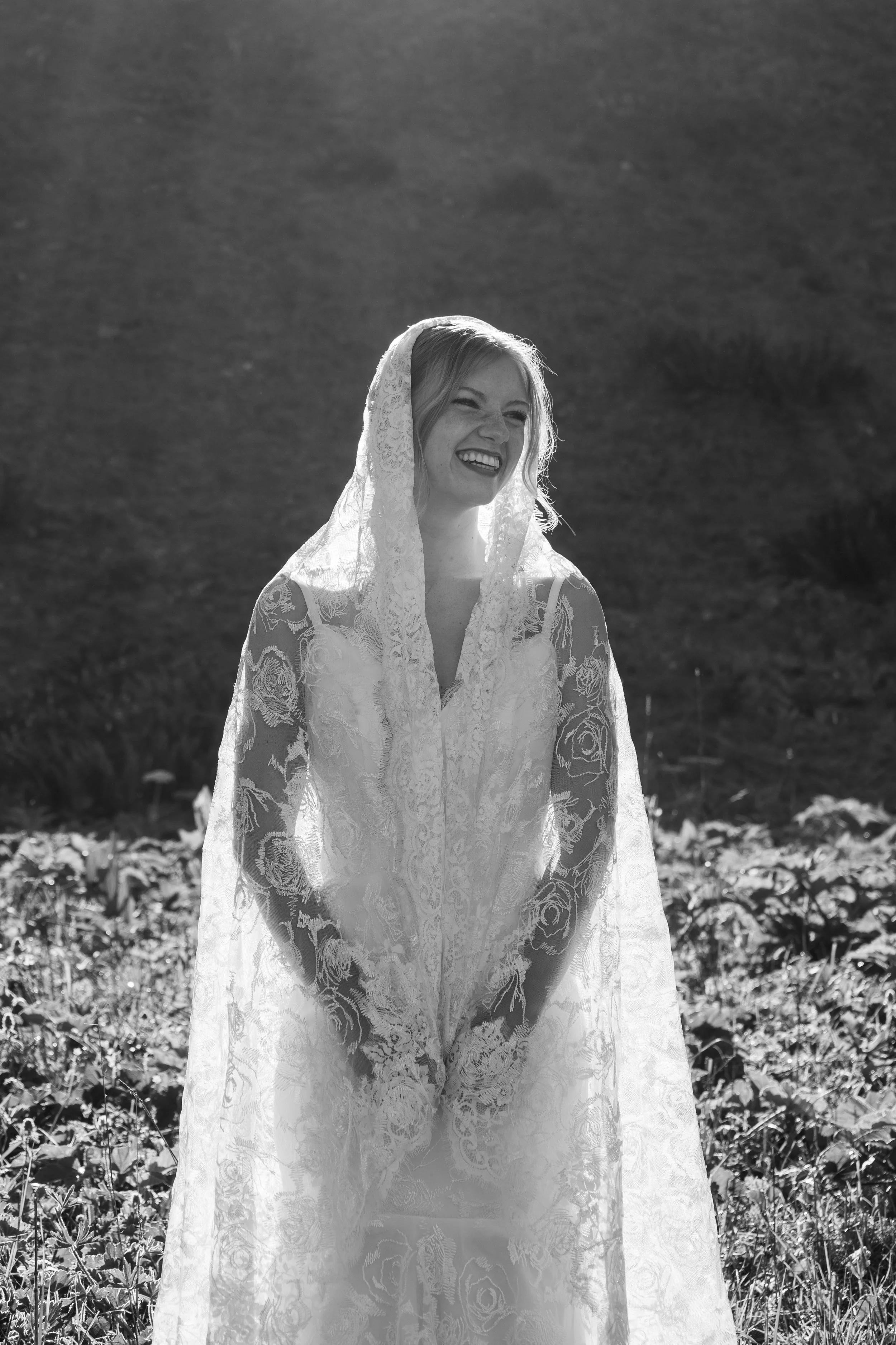 Black and white photo of a woman in lace wedding dress outdoors, smiling and wearing a lace veil.