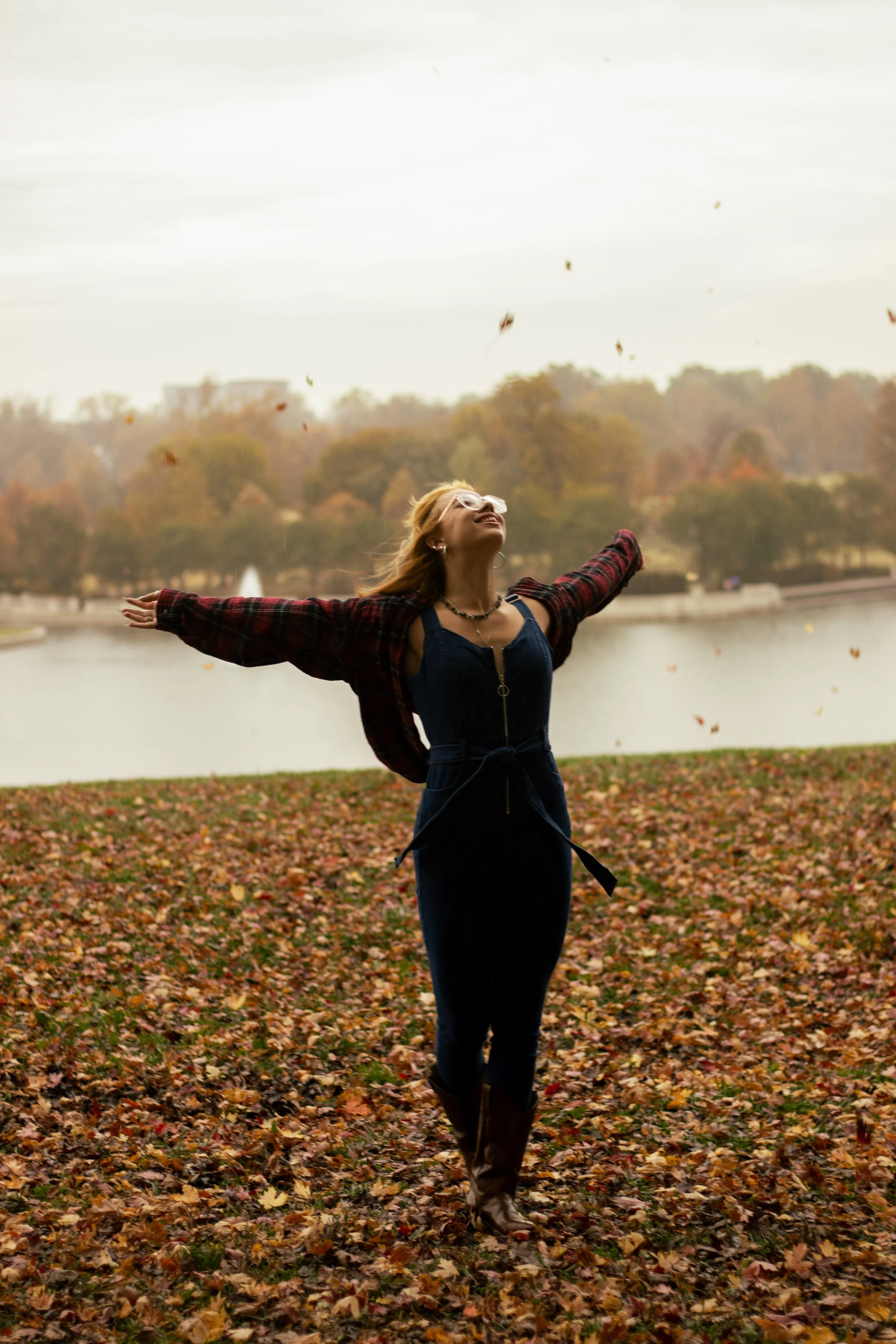 A woman in a dark jumpsuit, plaid jacket, and boots enjoying fall leaves at a park with a lake in the background.