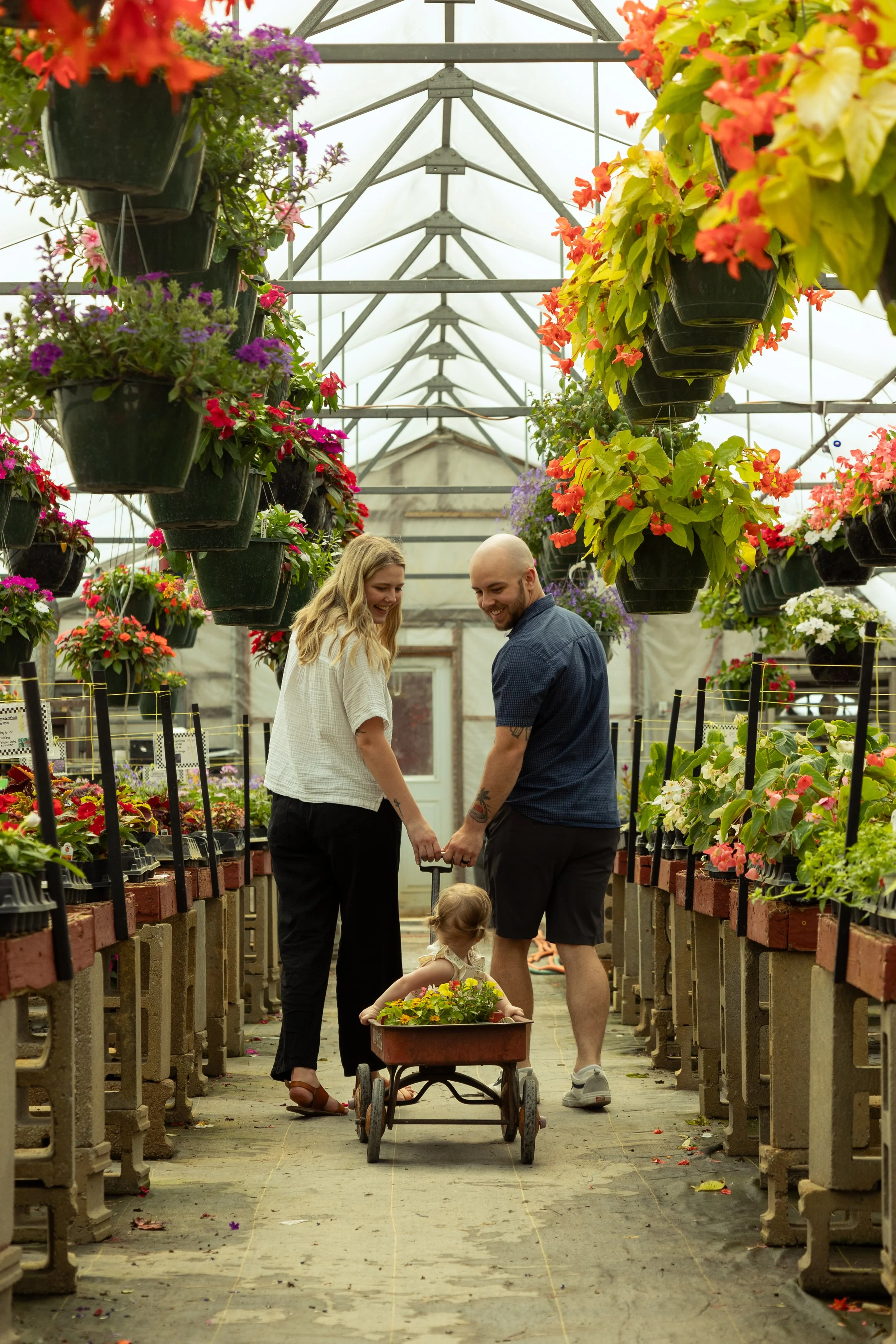 A happy family of three, with a young child in a wagon, holding hands in a greenhouse filled with colorful flowers.