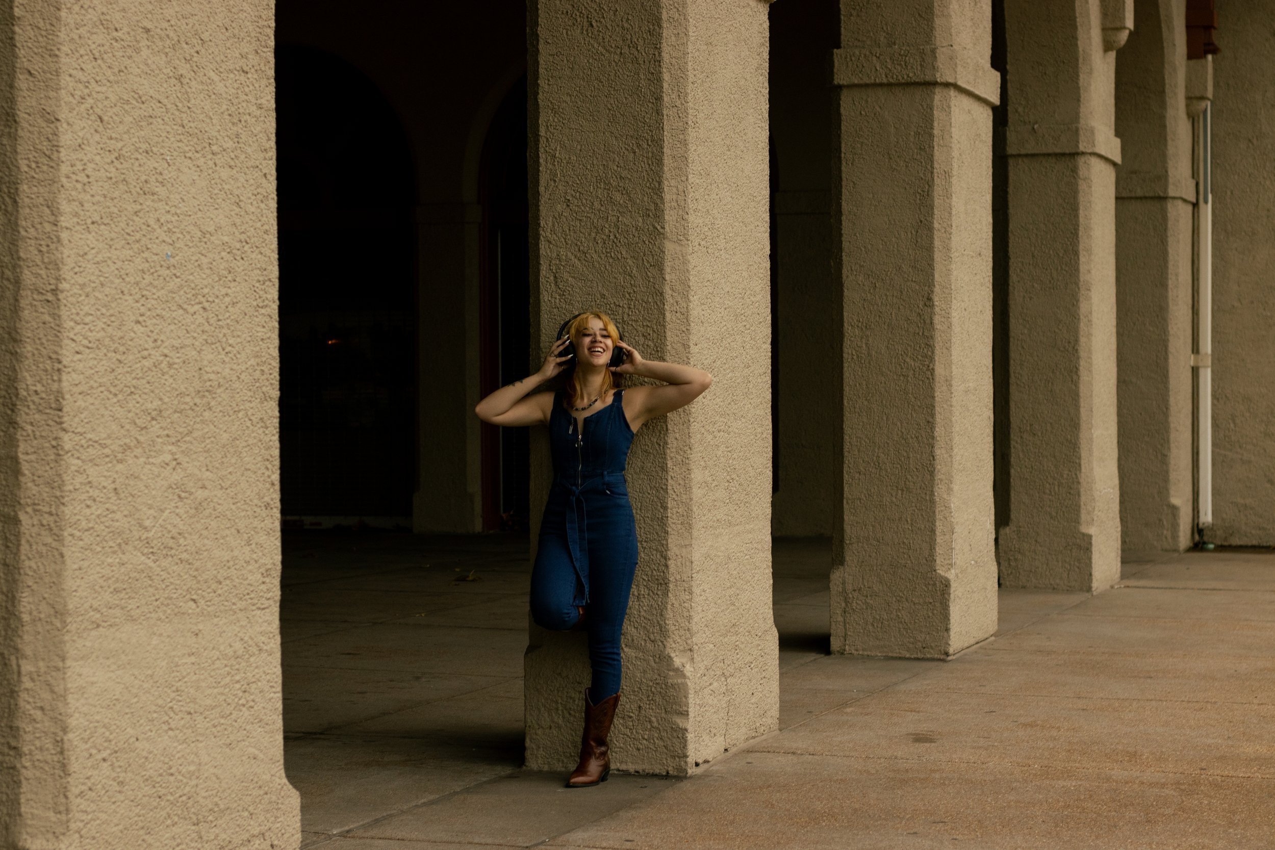A young woman with blonde hair smiling and listening to music with headphones, standing against a beige textured wall in an outdoor setting at night.