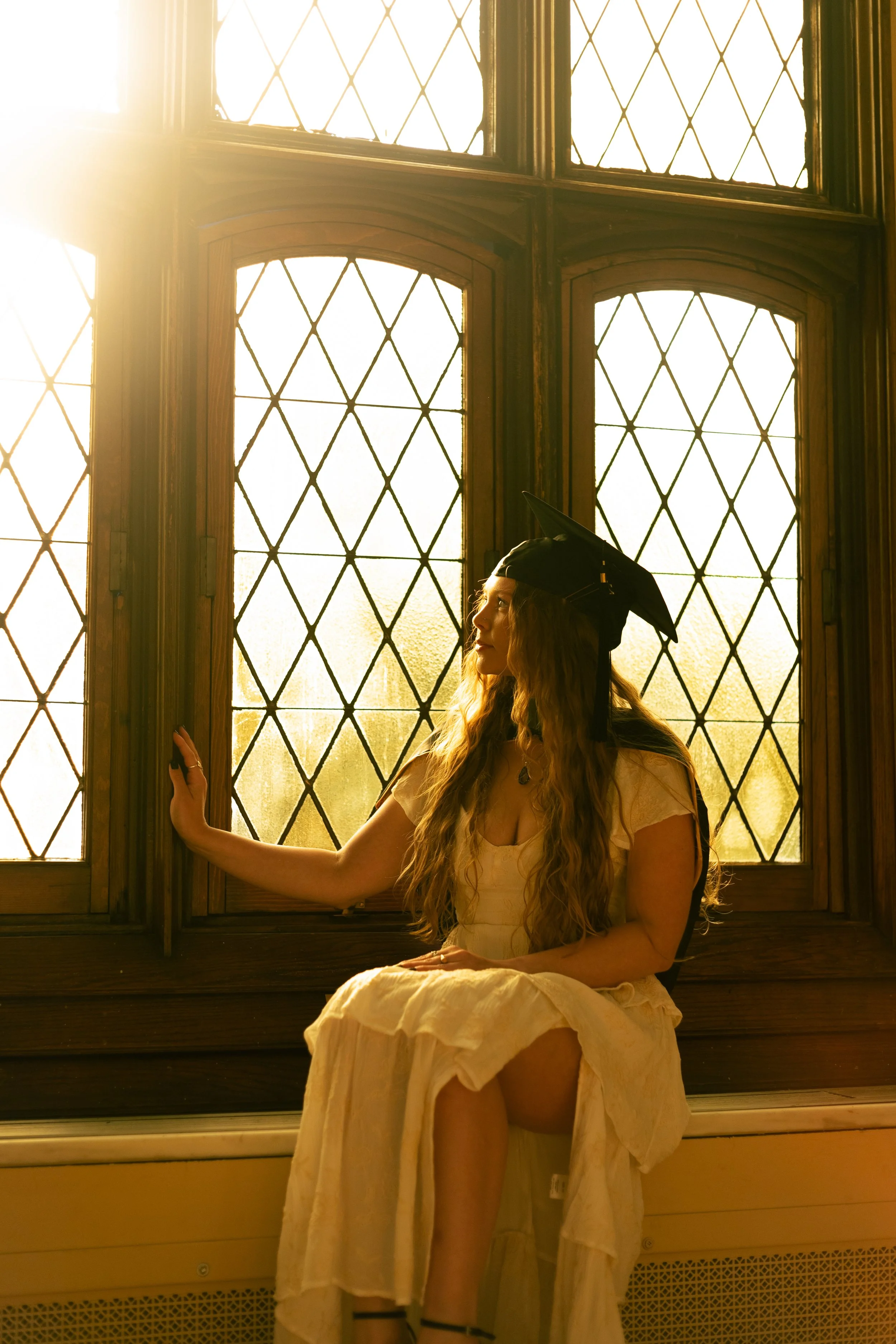 A young woman with long, curly hair wearing a graduation cap, sitting on a ledge by a large stained glass window with diamond patterns, illuminated by warm sunlight.