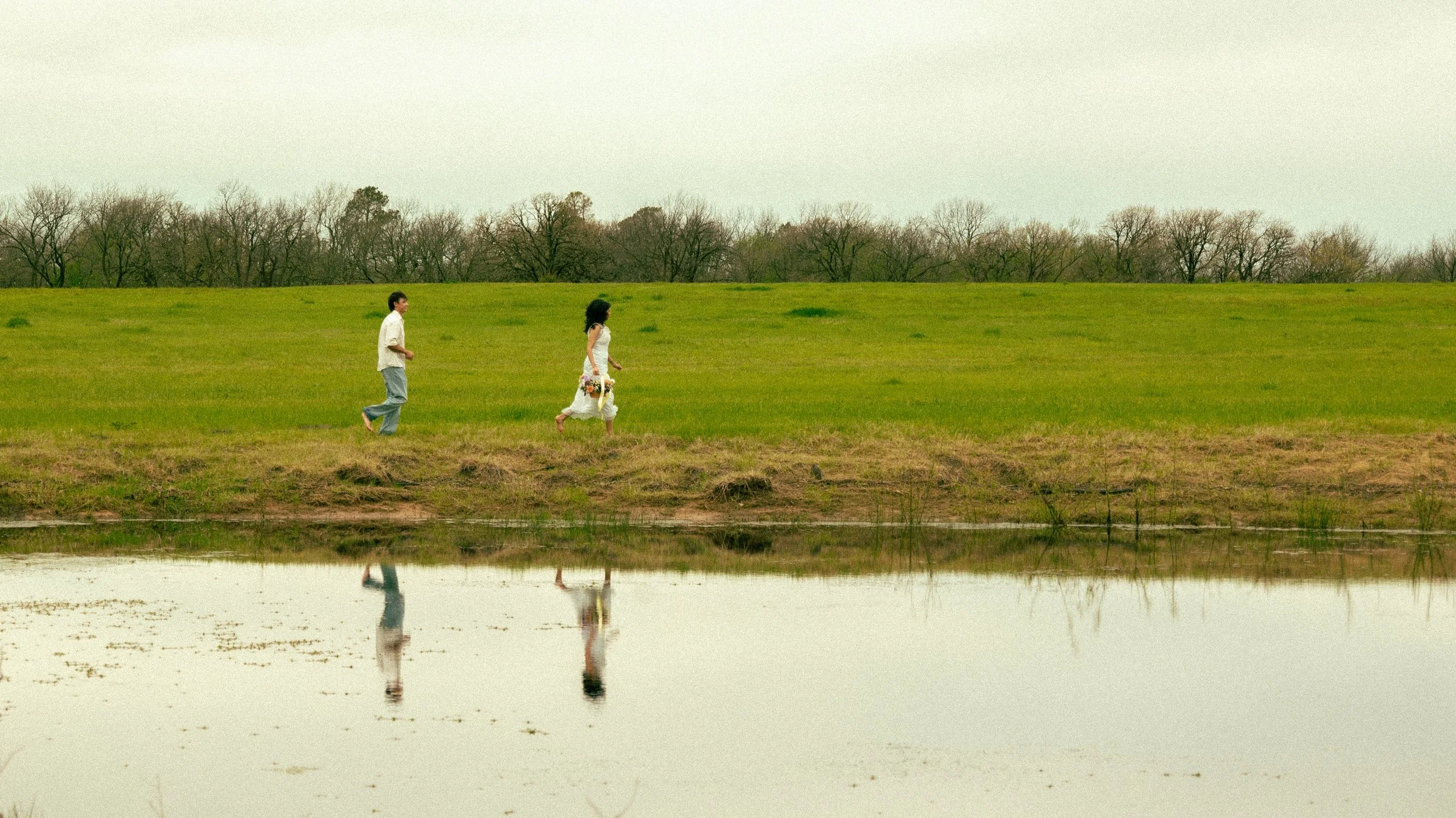 A couple walking along a grassy riverbank during daytime, reflected in the calm water.