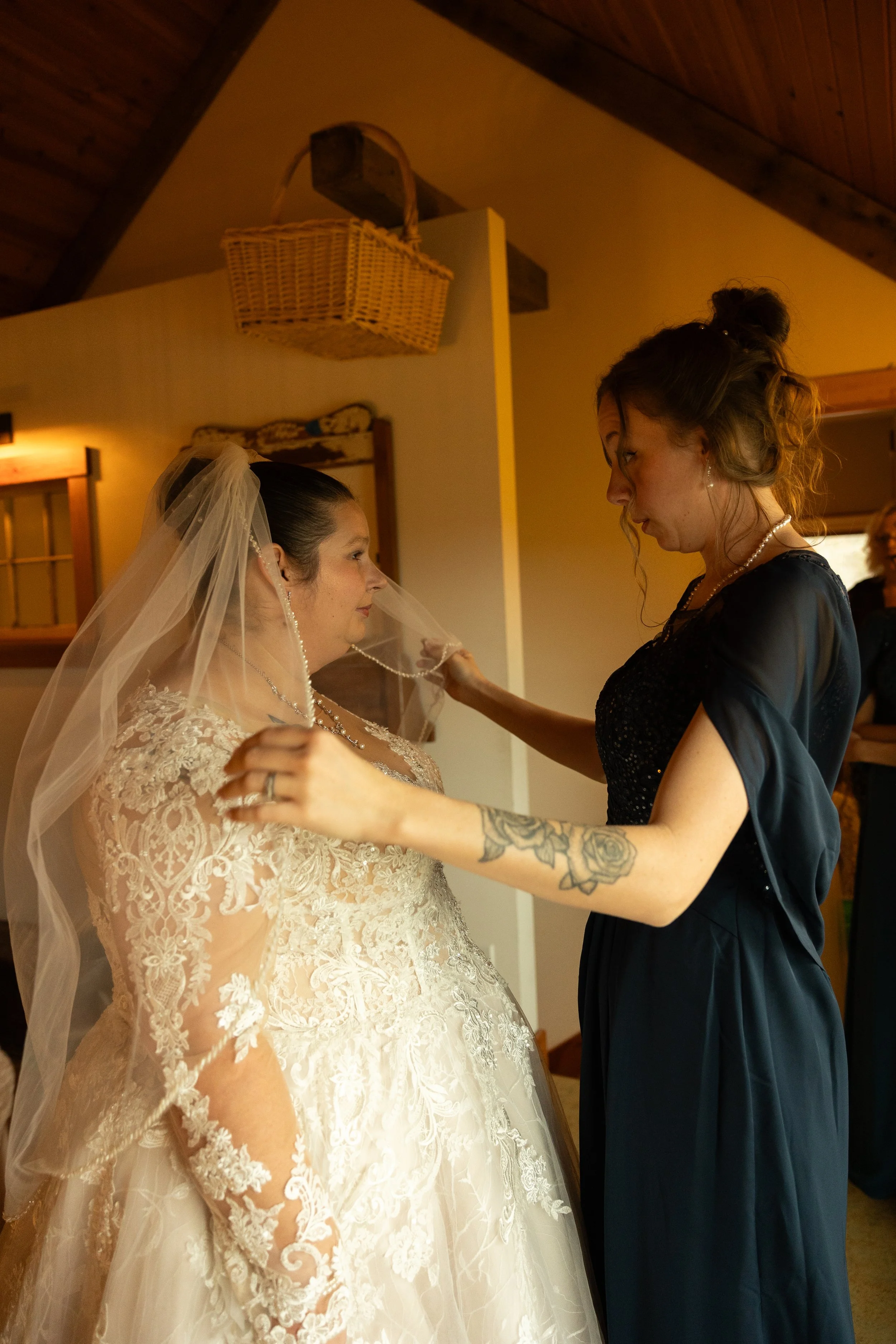 A bride in a lace wedding dress and veil is being assisted by a woman in a black dress as she puts on a necklace.