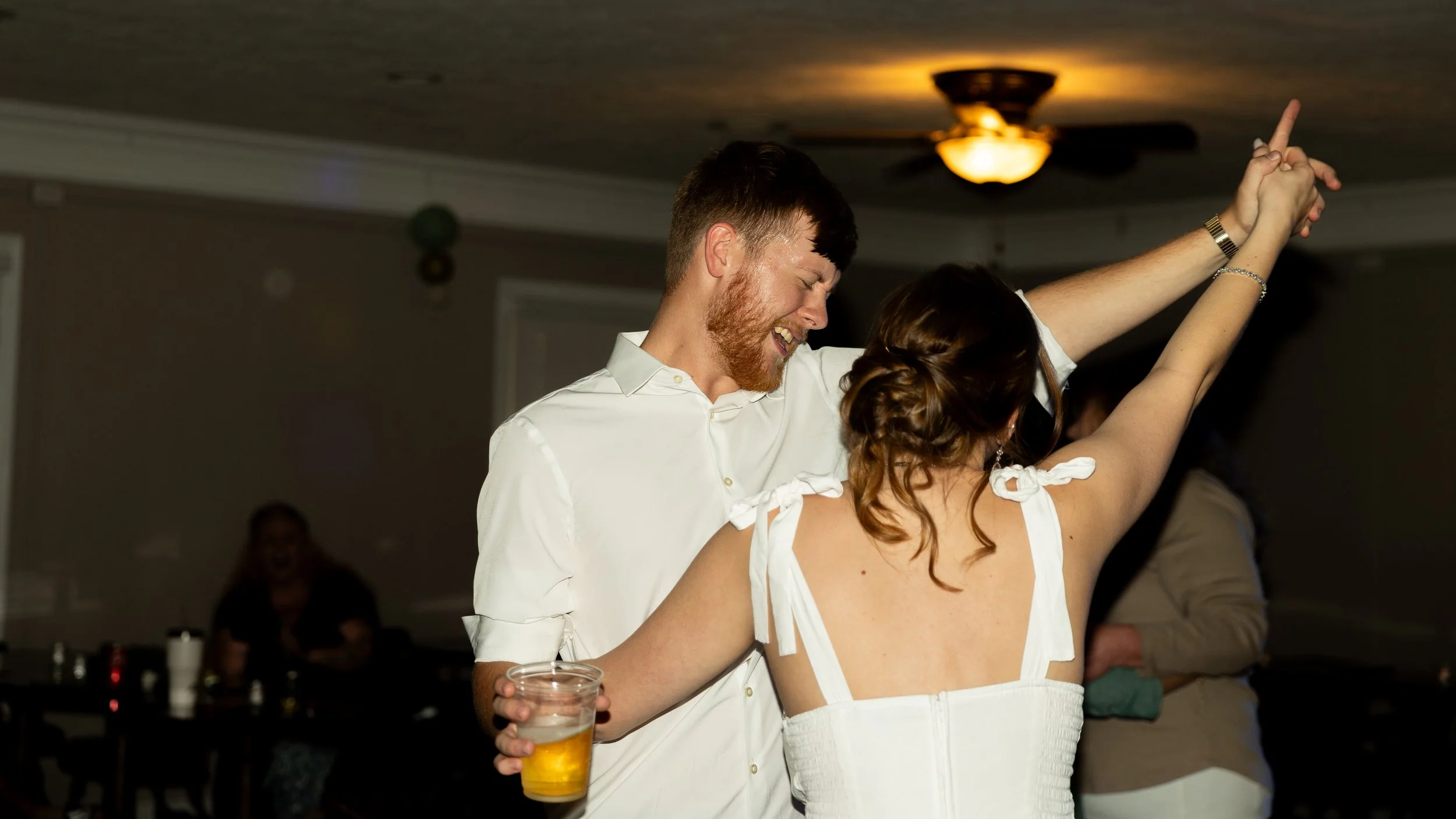A man and a woman dancing cheerfully at a party, with the man holding a drink and smiling, and the woman raising her arms in celebration.